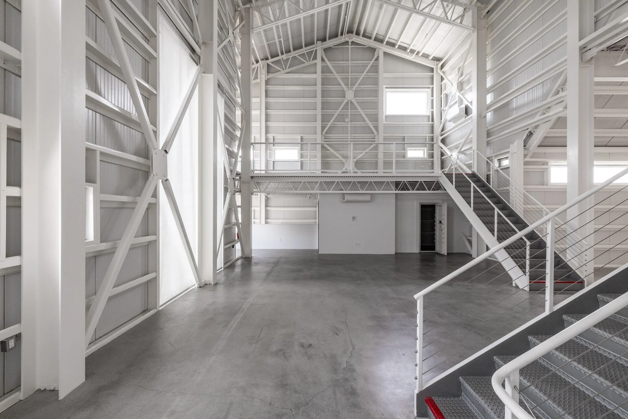 Empty industrial warehouse with white steel beams, stairs, metal railings, and concrete floor.