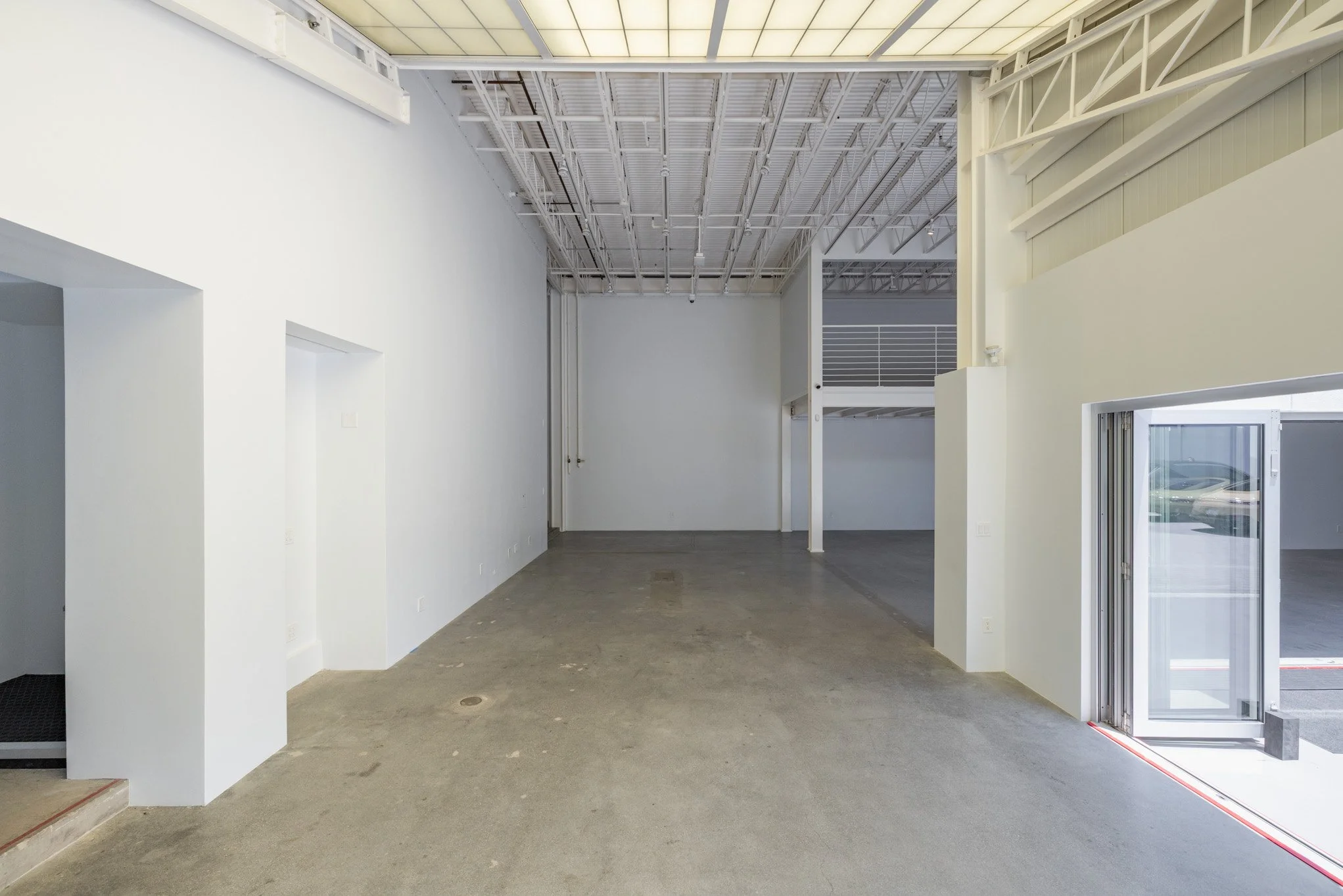 Empty commercial space with white walls, concrete floors, high ceiling with exposed beams, and a partially open glass door leading outside.