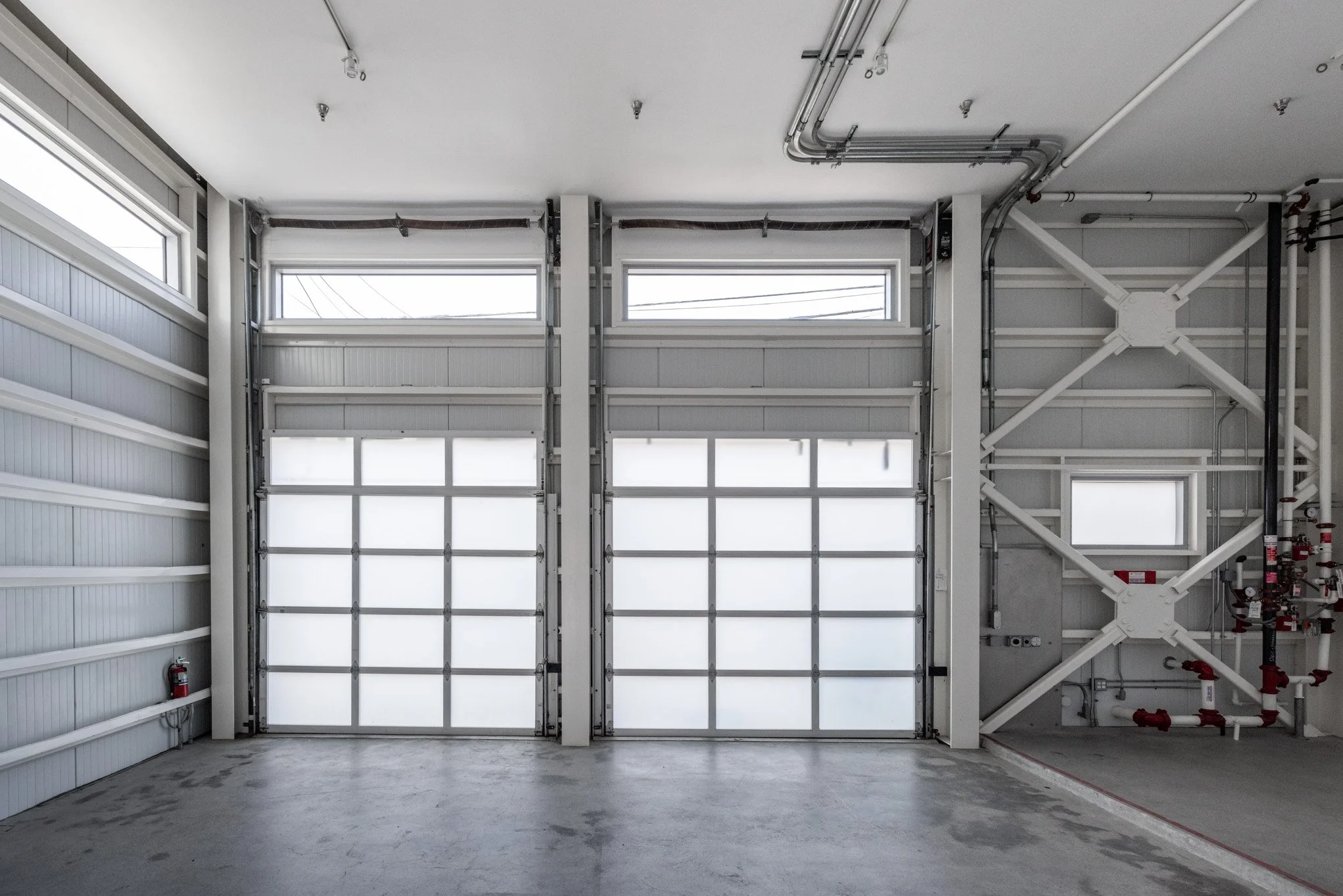 Empty garage with faux frosted glass doors, small side window, and visible industrial hardware and piping.