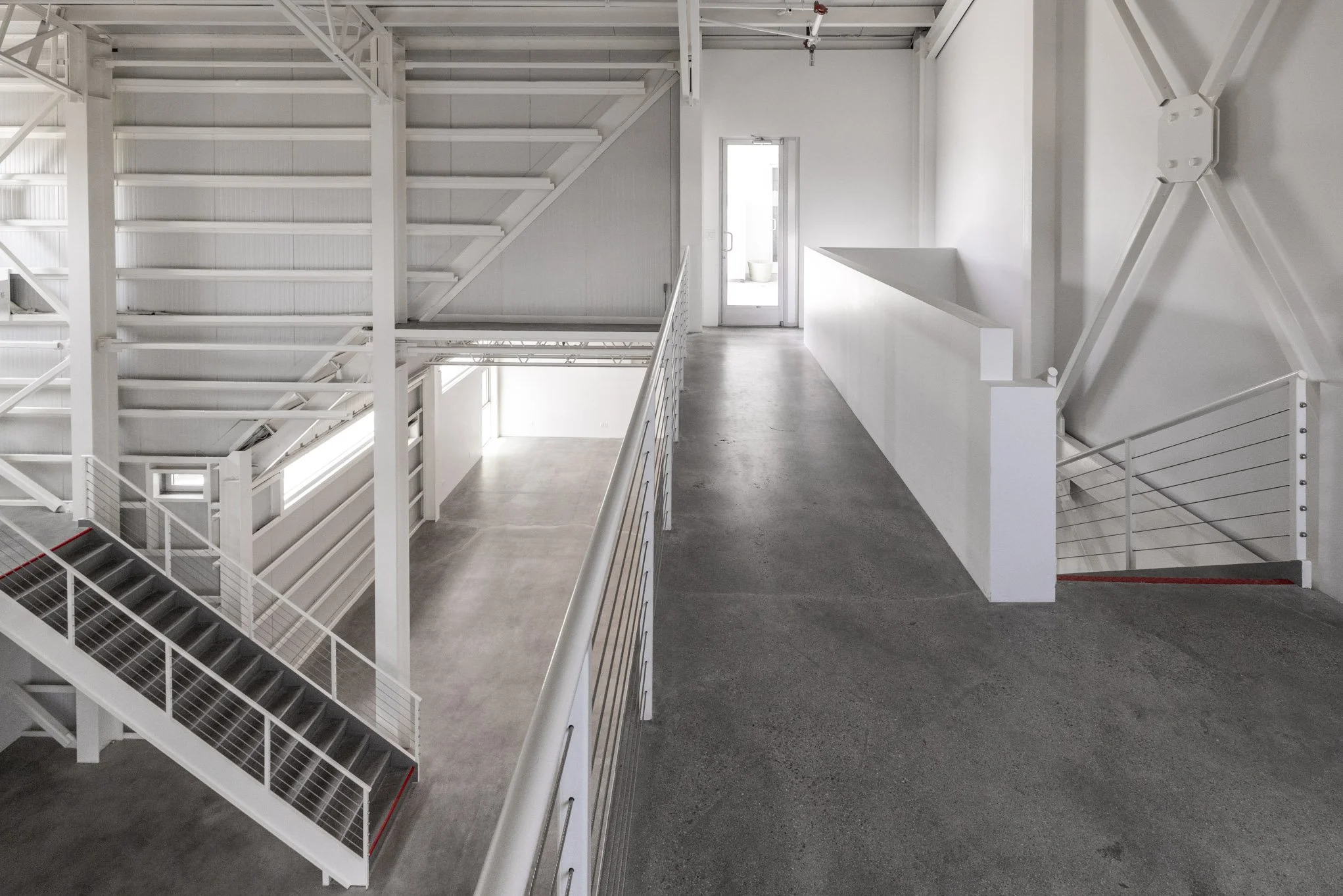 Empty modern interior space with white walls, metal staircases, and a concrete floor, featuring an upper level walkway with railing and a door at the end.