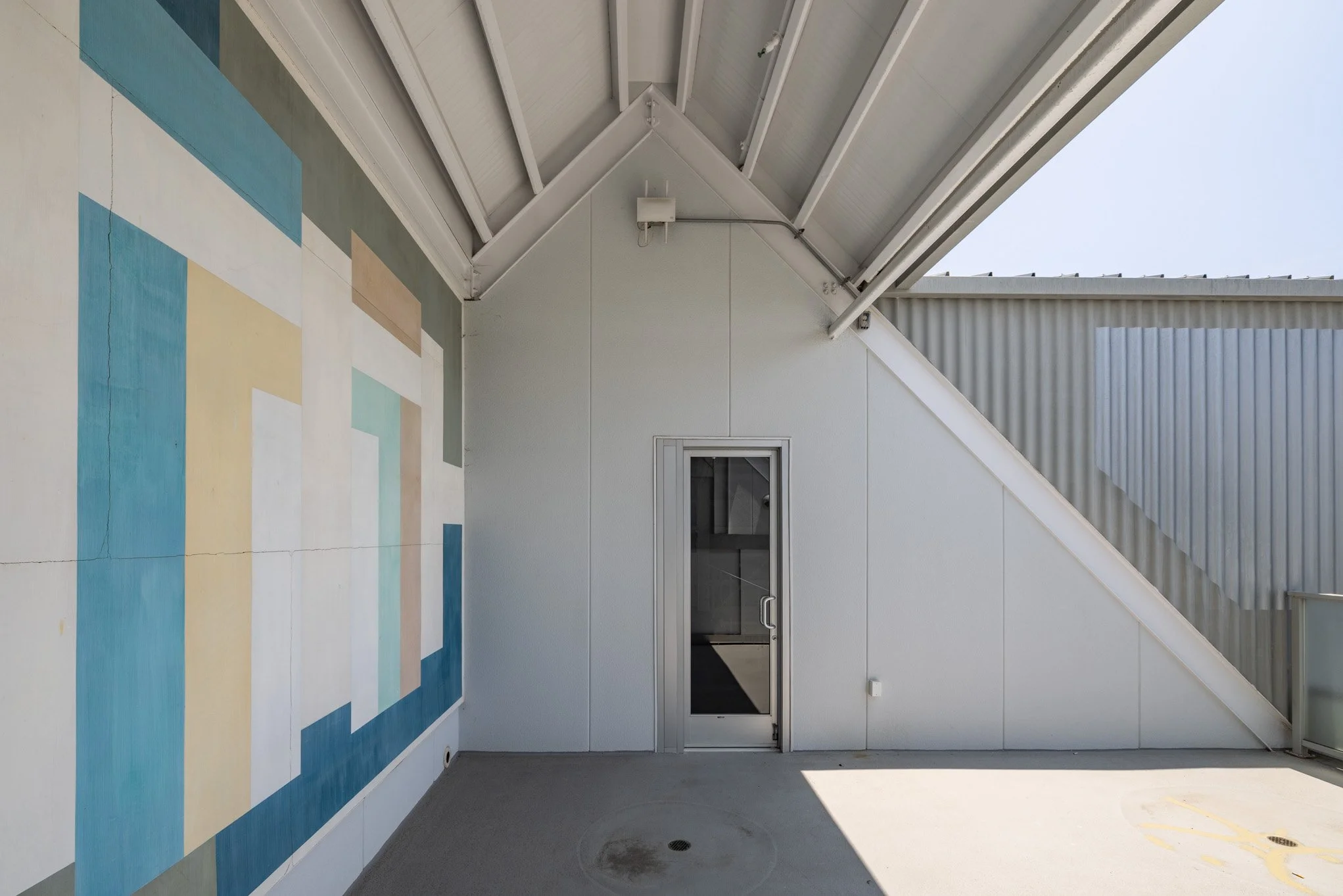 Empty rooftop patio with painted geometric mural on left wall, metal roof overhang, small door on back wall, and sunny sky overhead.