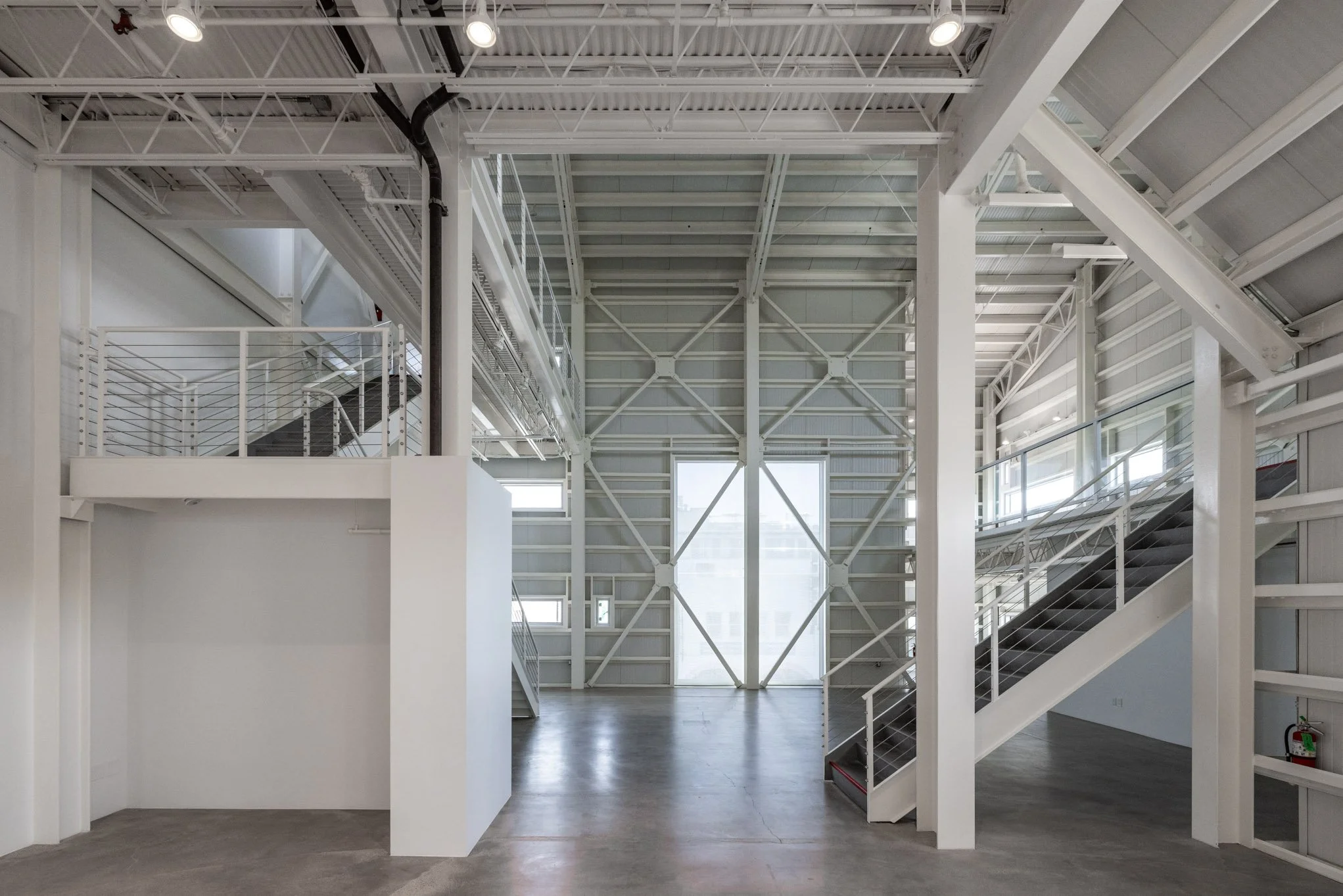 Empty industrial loft with white walls, metal staircases, and large windows, featuring an open floor plan and exposed ceiling beams.