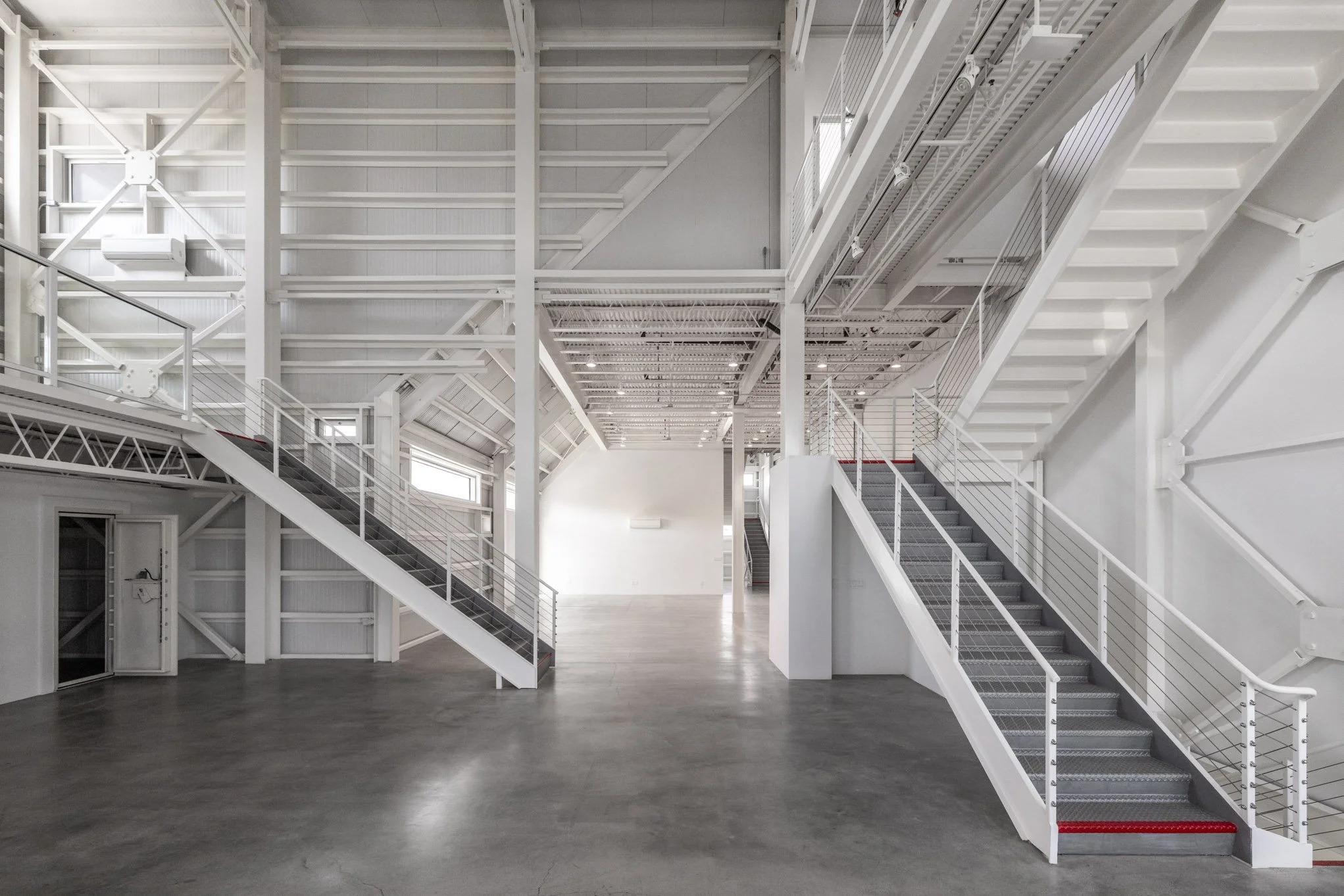 Empty industrial-style interior with white metal stairs, railings, and high ceiling, large open floor space, and natural light from windows.