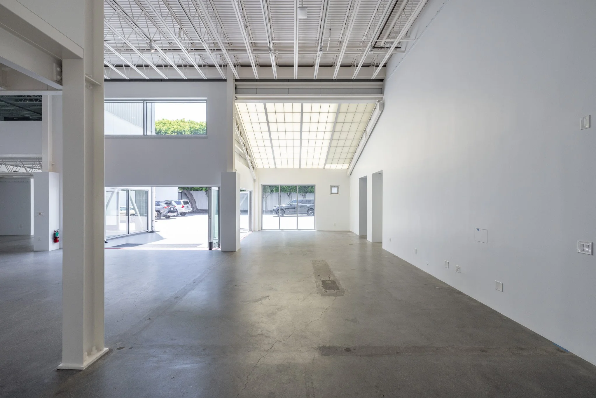 Empty commercial space with white walls, concrete floor, large windows, and a high ceiling with exposed white beams.