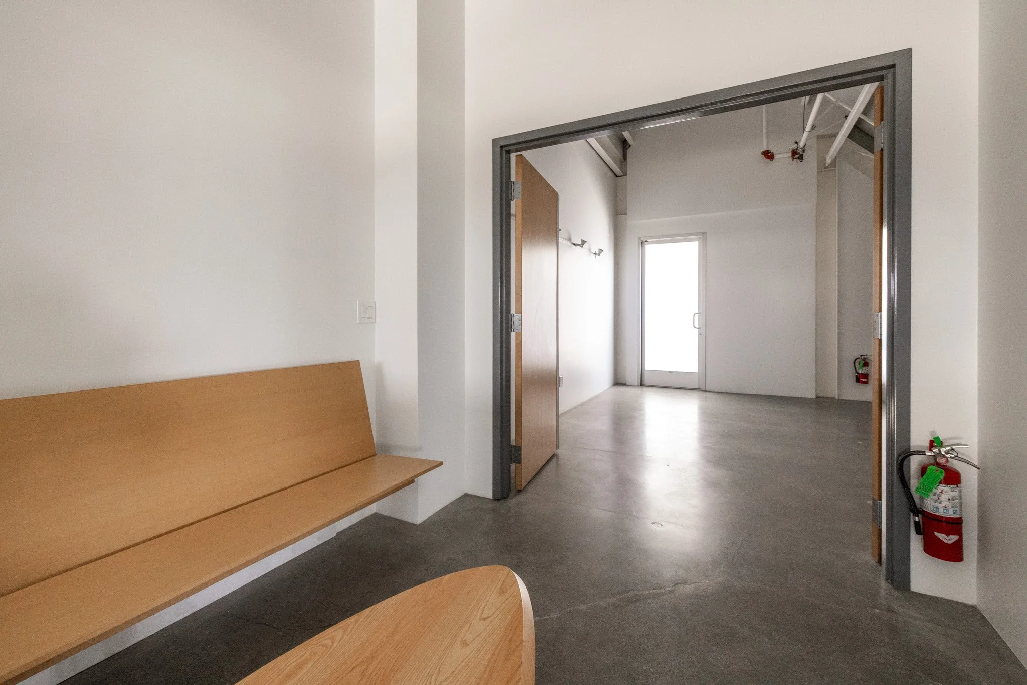 Interior of an empty room with a wooden bench, gray floor, white walls, and a doorway leading to another room with a glass door and window.