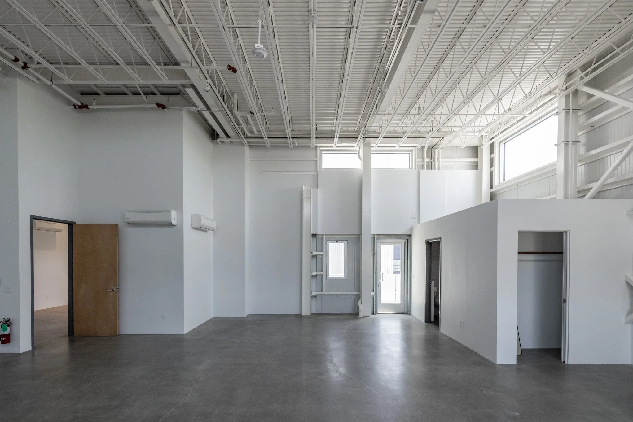 Empty interior space with white walls, concrete floor, and high ceiling with exposed metal beams and skylights, featuring multiple small rooms with doors.