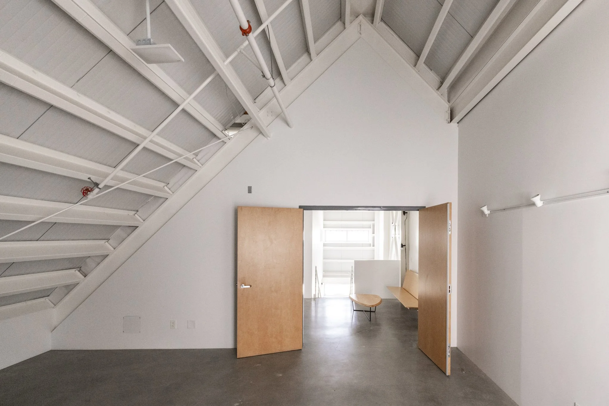 Empty modern interior space with white walls, wooden doors, stairs on the left, and a seating area with a bench and a small table visible through open doors.