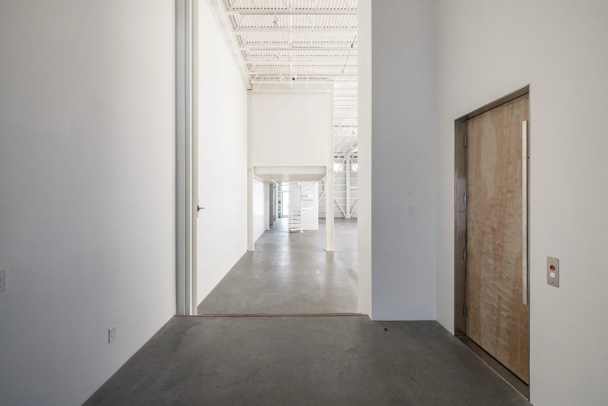 Empty, white-walled interior space with concrete flooring, an elevator door on the right, and a hallway with a staircase in the distance.