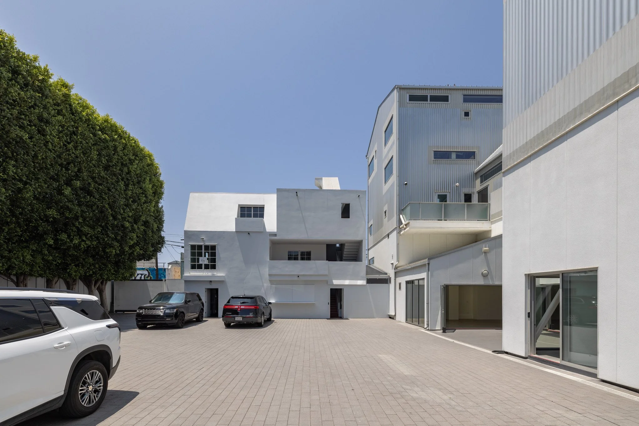 Modern white and gray multi-story buildings in a paved courtyard with parked cars, a large green tree on the left, and a clear blue sky.