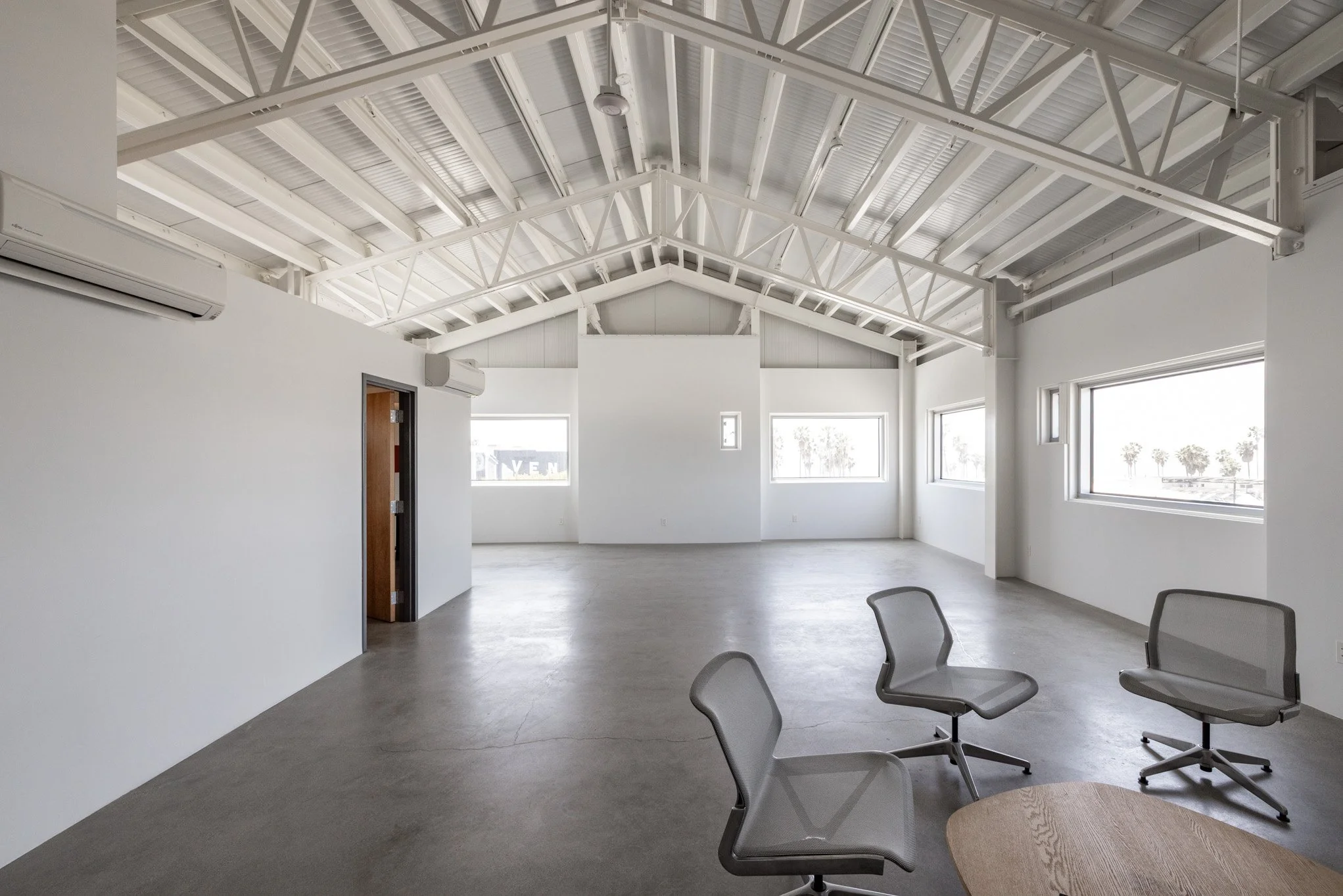 Empty modern open office space with white walls, concrete floor, large windows, exposed ceiling beams, and a few office chairs around a round table
