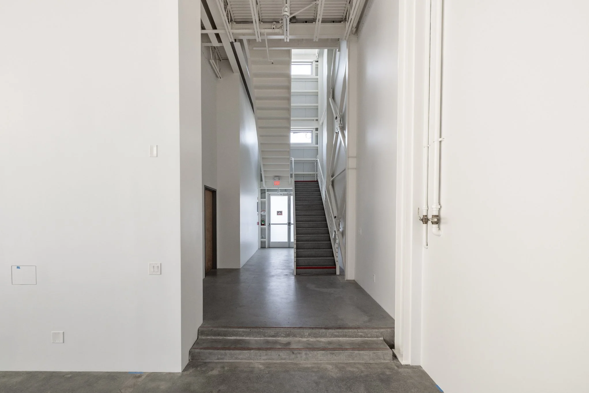 Interior view of a modern building with white walls, a staircase leading to an upper level, and a glass door at the back with windows allowing natural light.
