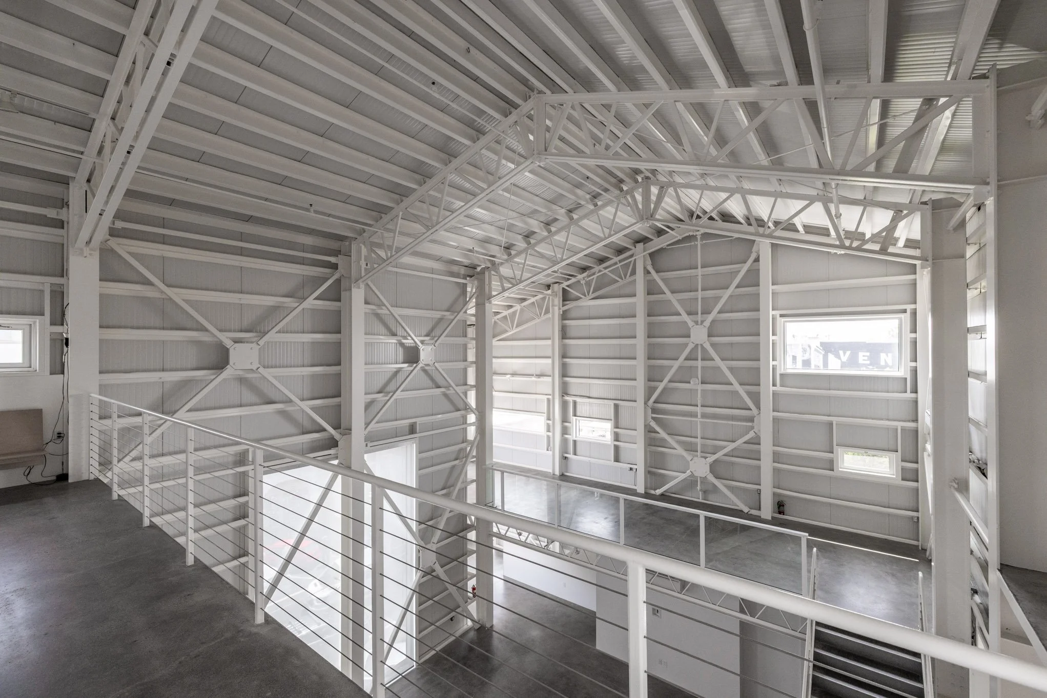 Interior view of an industrial or warehouse building with white steel framing, high ceiling, and multiple small windows, with a mezzanine area and staircase.