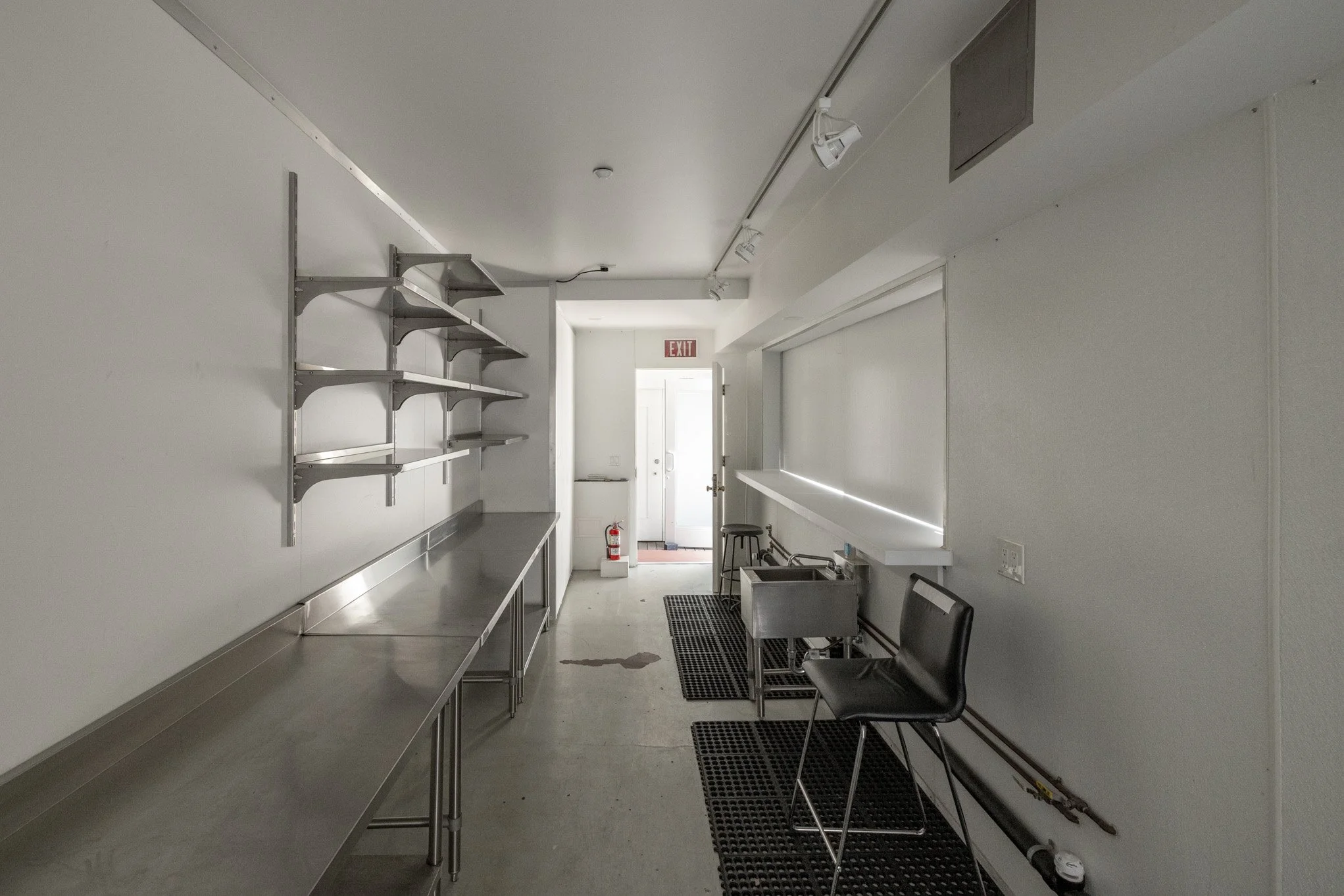 Empty commercial kitchen with stainless steel counter and shelving, black chairs, wall-mounted sink, and entrance door in the background.