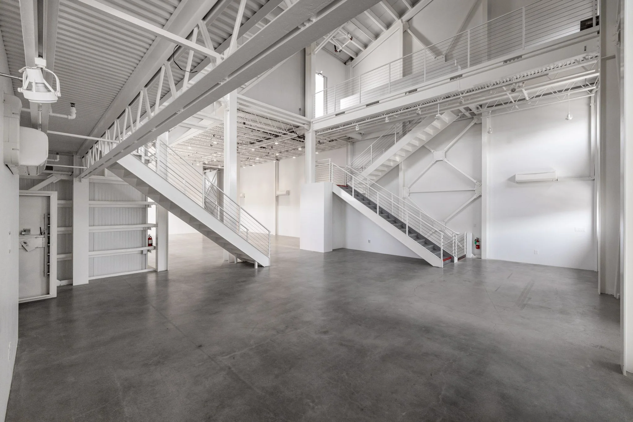 Empty industrial or warehouse space with white walls, concrete floor, and metal stairs leading to an upper level.