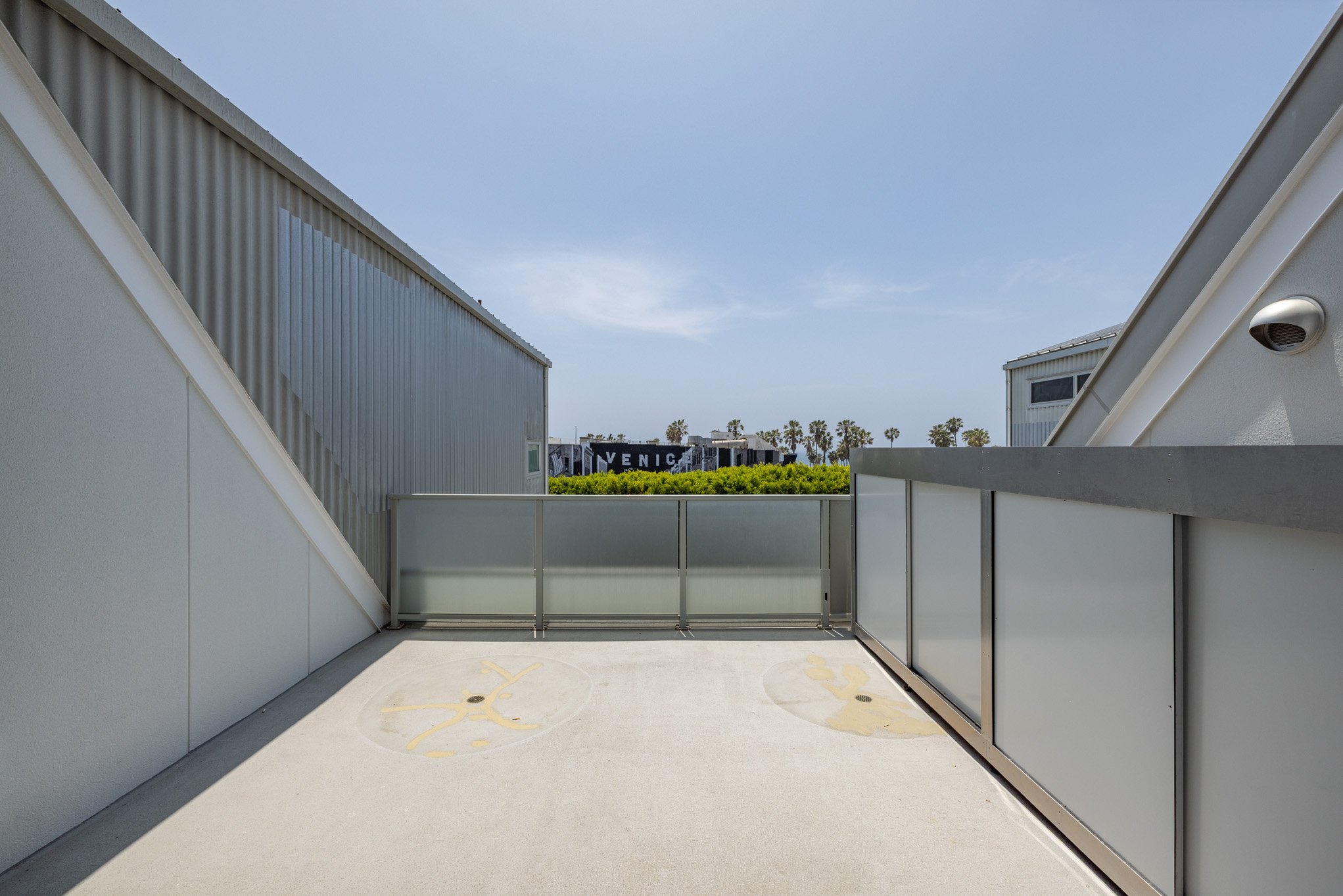 Empty rooftop terrace with frosted glass railing and two circular marked spots on the concrete floor, overlooking palm trees and a blue sky.