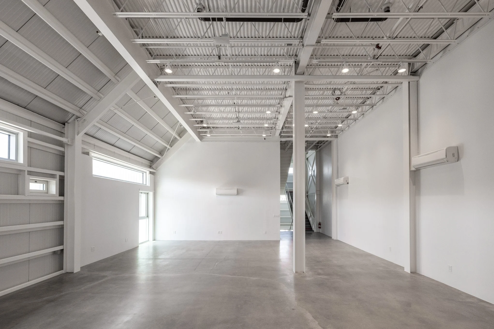 Empty, spacious interior with white walls, polished gray concrete floor, high vaulted ceiling with exposed metal beams, and multiple windows, suggesting an industrial or commercial space.