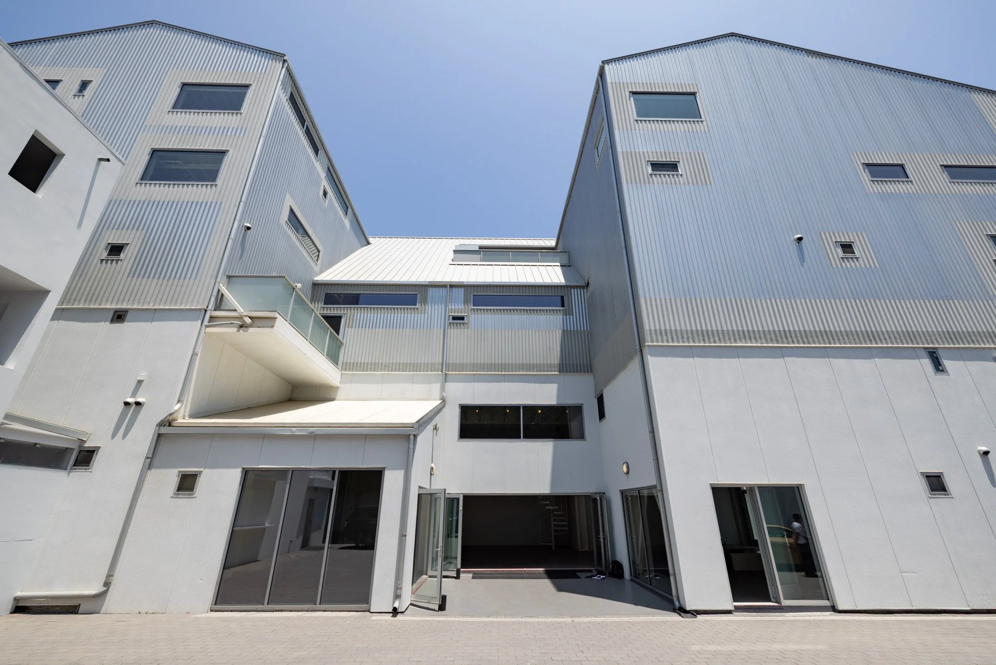 Modern multi-story building with a mix of white concrete and metal siding, glass doors and windows, and an open garage area at the bottom, under a clear blue sky.