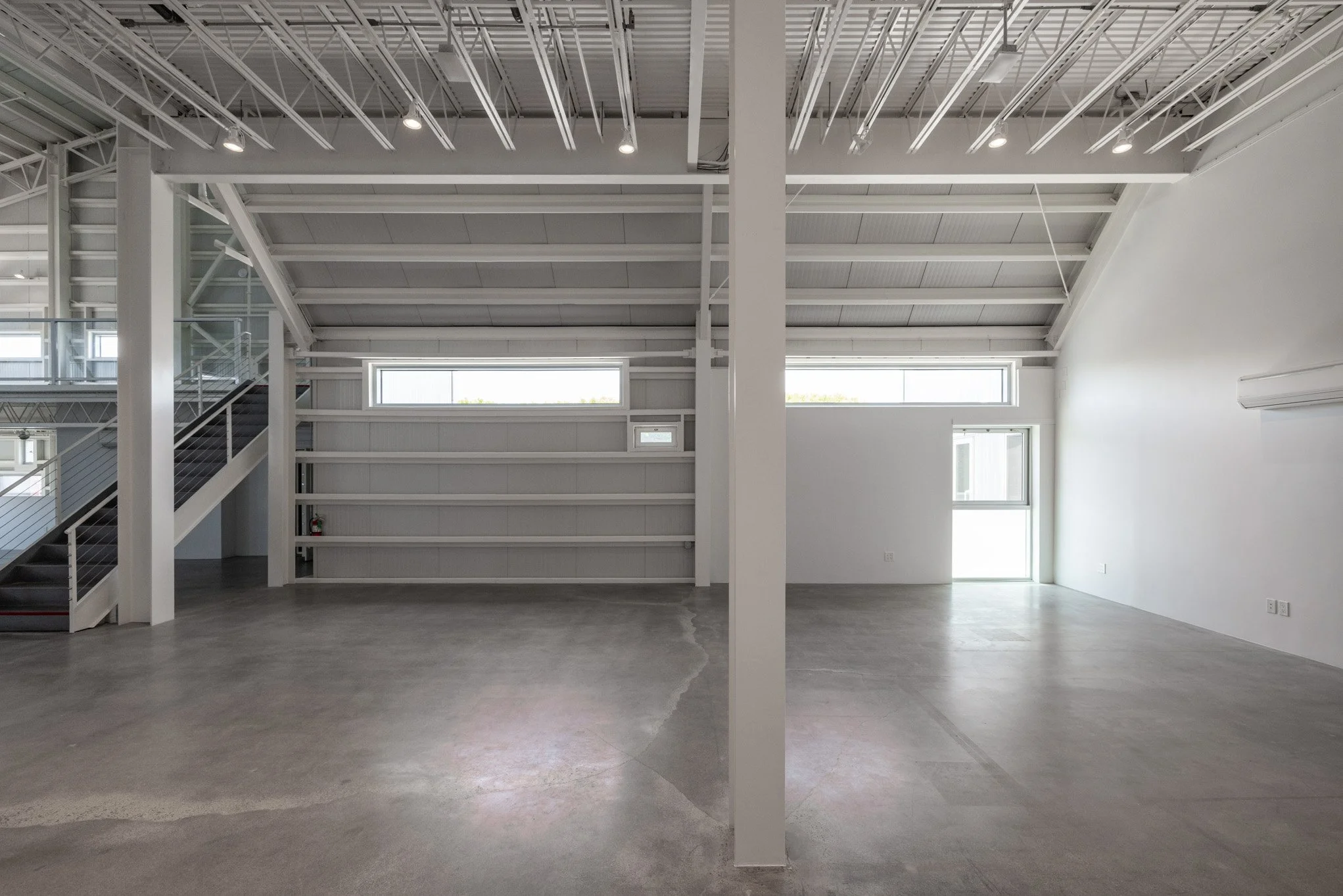 Empty industrial-style space with high ceilings, metal beams, white walls, concrete floors, interior staircase, and large windows.