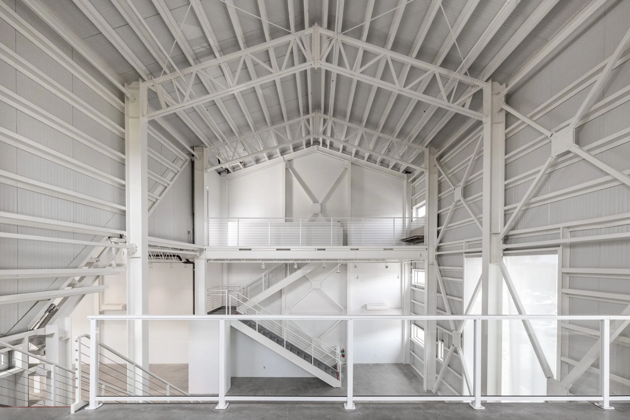 Interior view of a large, modern, white industrial warehouse with high vaulted ceiling and metal support beams, multiple small windows, and a staircase leading to an upper mezzanine level.