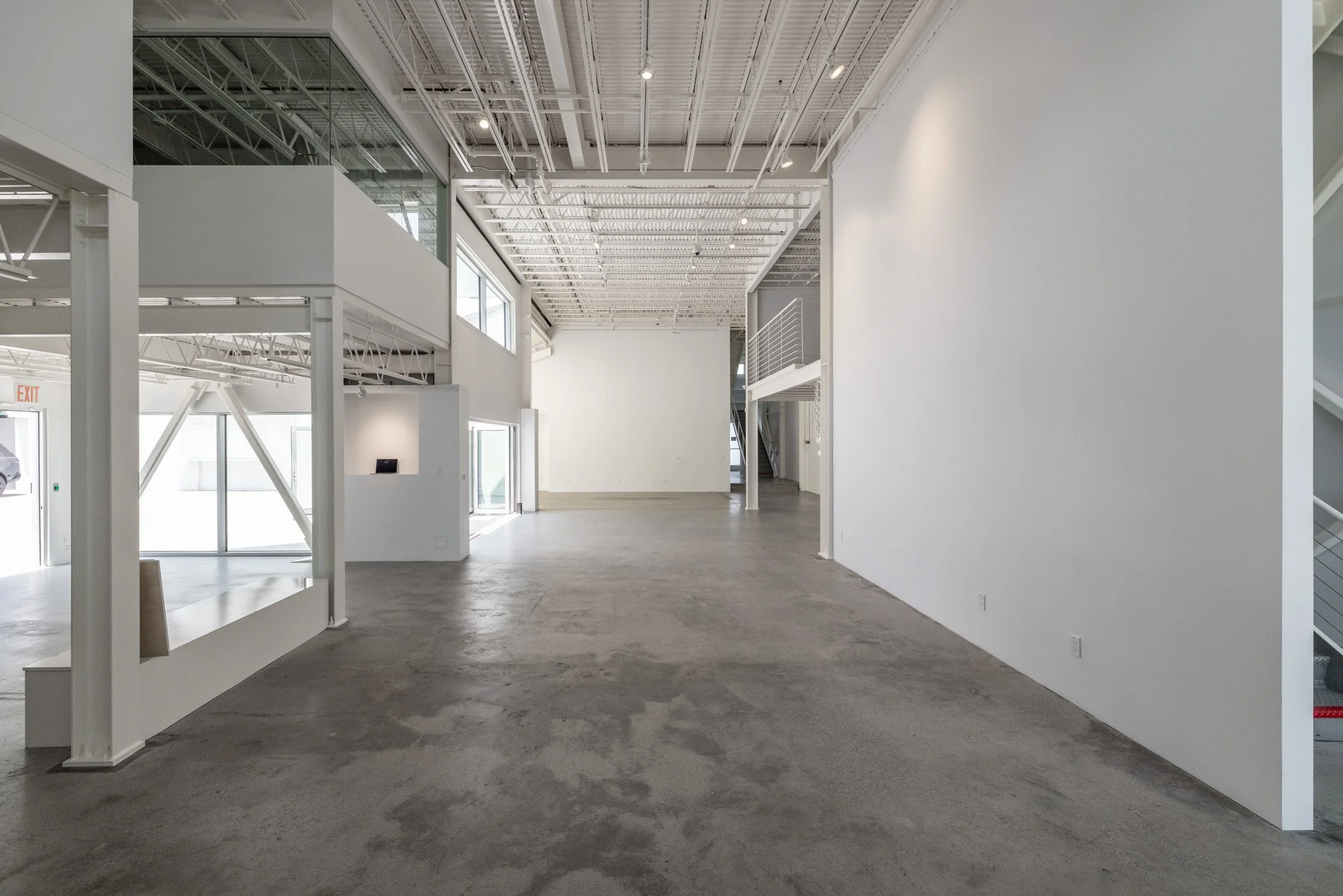Empty modern commercial interior with high ceilings, exposed structural elements, white walls, and large windows.