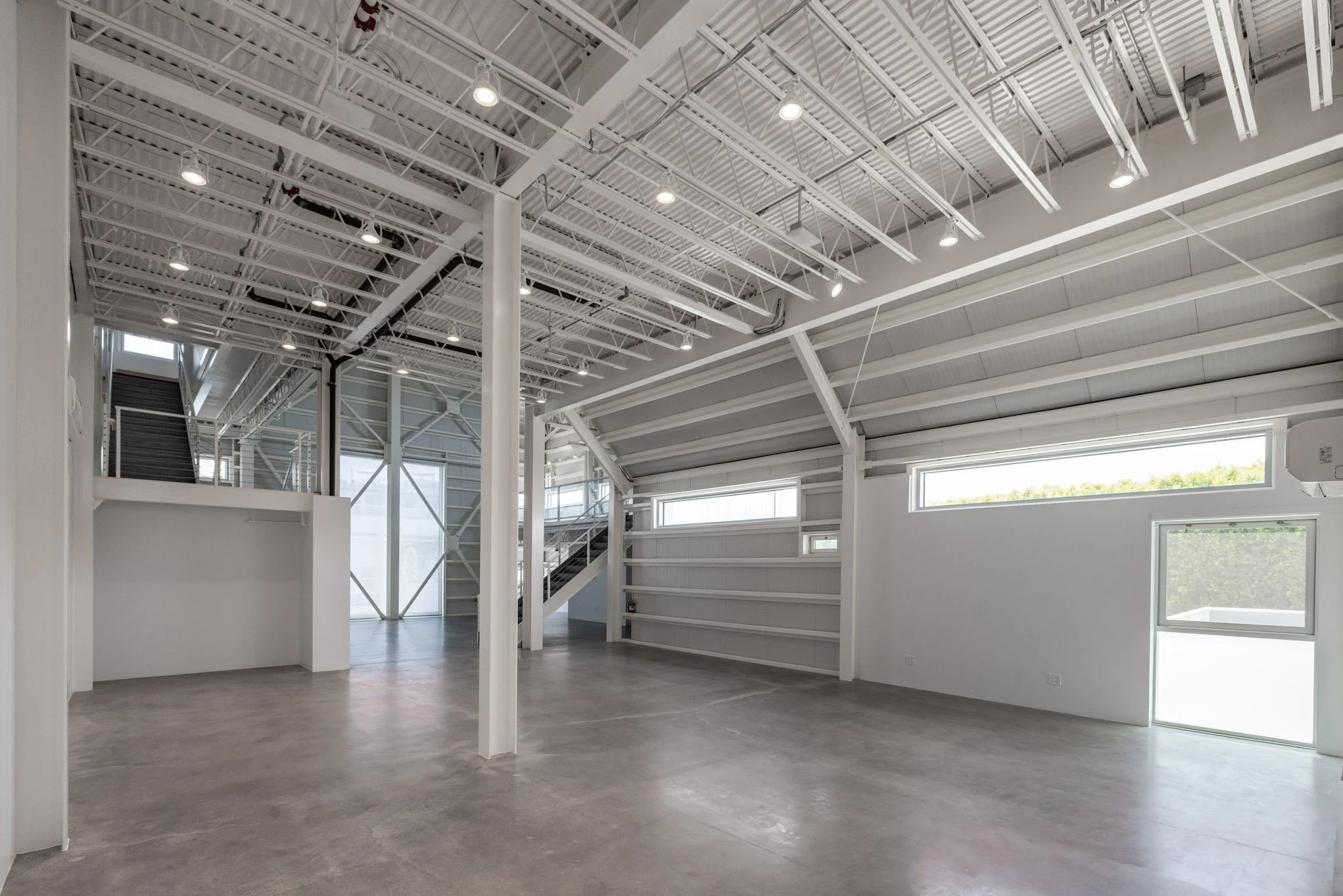 Empty interior space with high ceilings, large windows, and exposed ceiling beams, finished with concrete floors.
