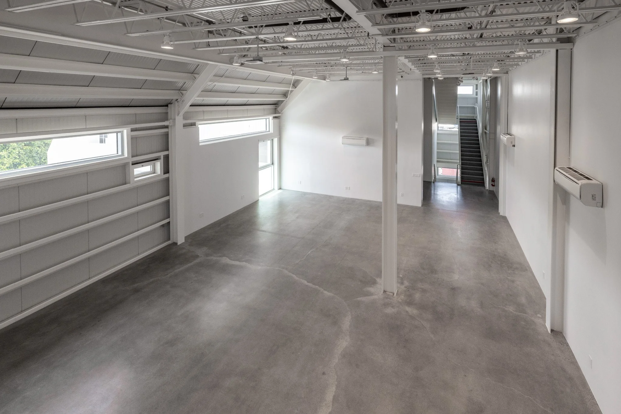 Empty industrial-style interior space with concrete floors, white walls, large windows, and exposed ceiling beams, featuring stairs at the back.