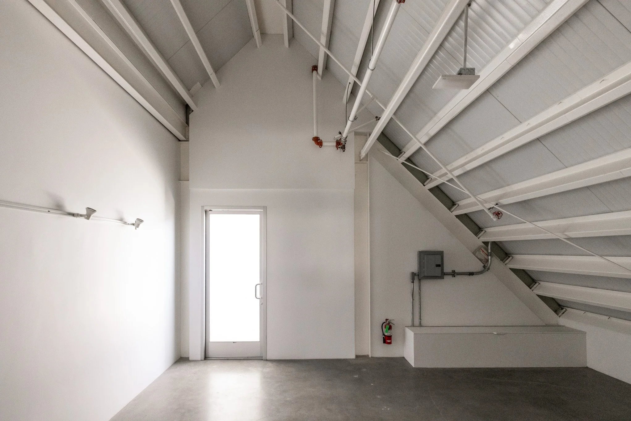 Empty room with white walls and ceiling, door at the back, and visible plumbing pipes with sprinkler system. Fire extinguisher mounted on the wall to the right of the door.
