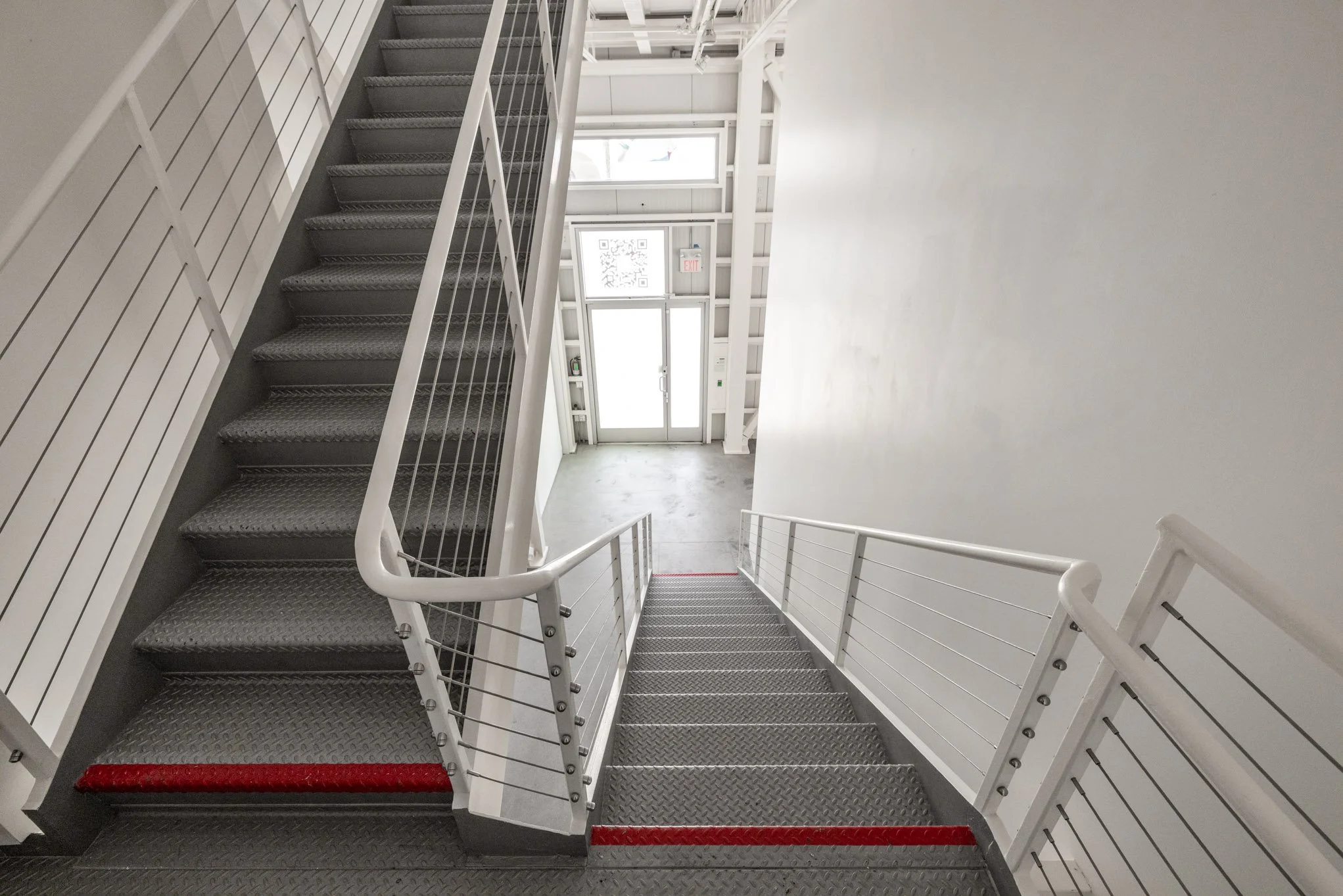 Interior view of a stairwell with gray metal stairs, white railings, and an exit door with a window at the bottom, allowing light into the space.