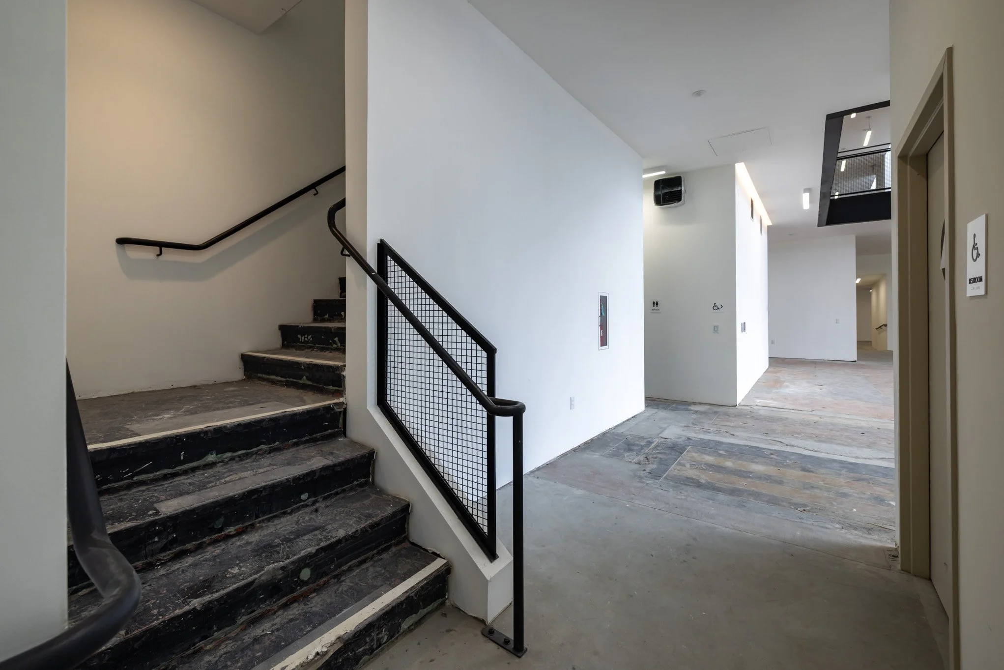 Interior view of a building in progress with stairs on the left, white walls, a modern black railing, and a spacious hallway with signs indicating accessible and restroom facilities.