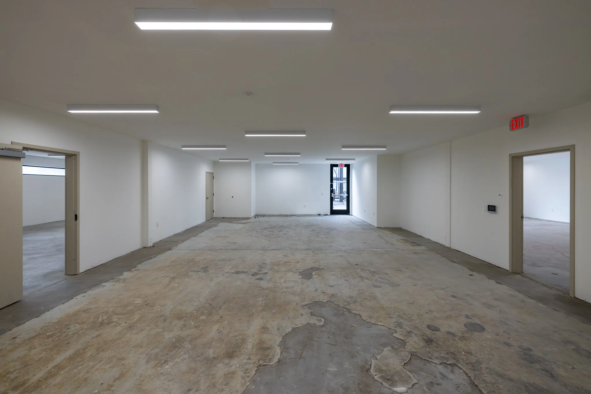 Empty commercial interior space with white walls, fluorescent ceiling lights, and an unfinished floor, with doorways leading to smaller rooms.