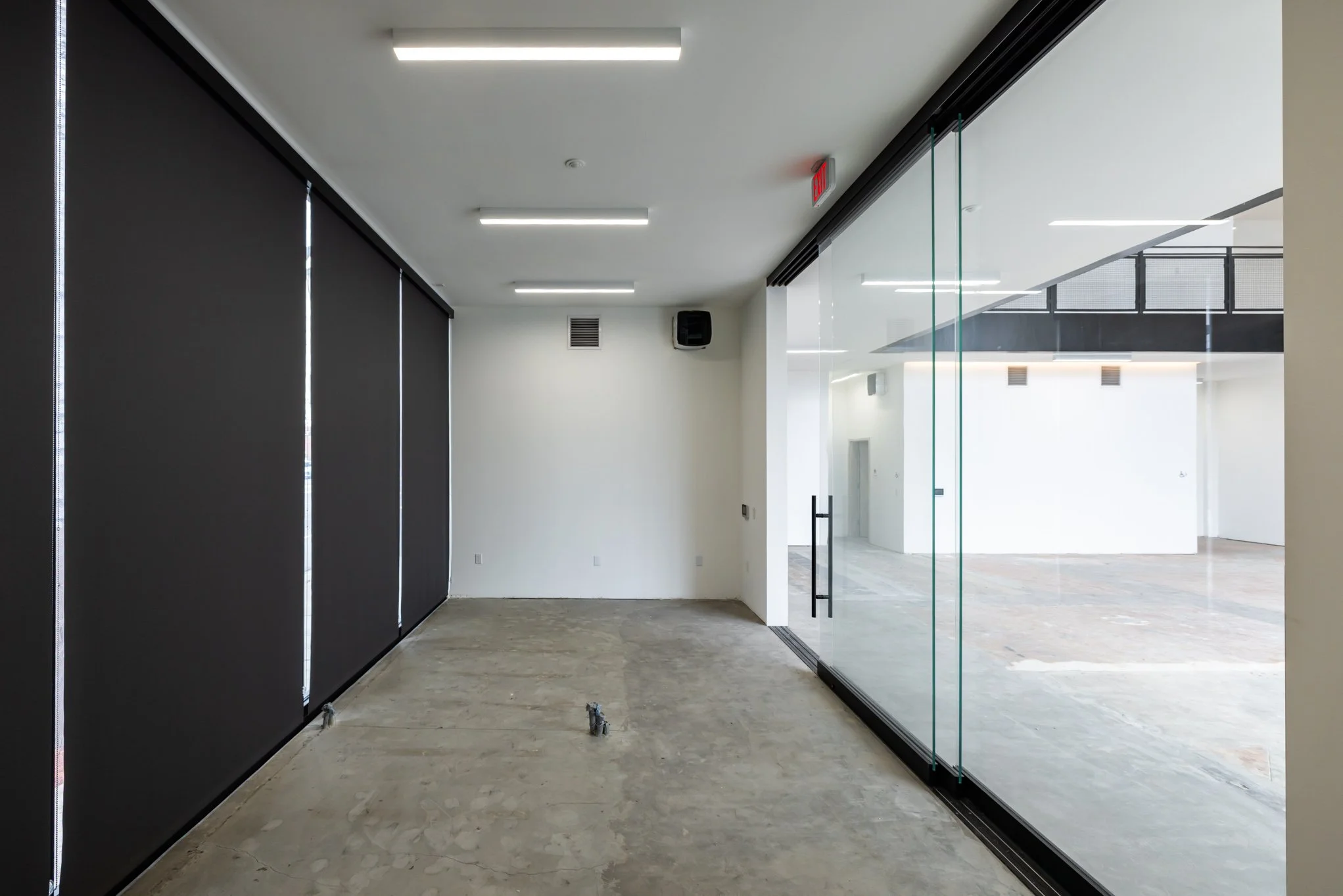 Empty modern office space with black roller blinds on the left, glass walls on the right, and unfinished concrete flooring.