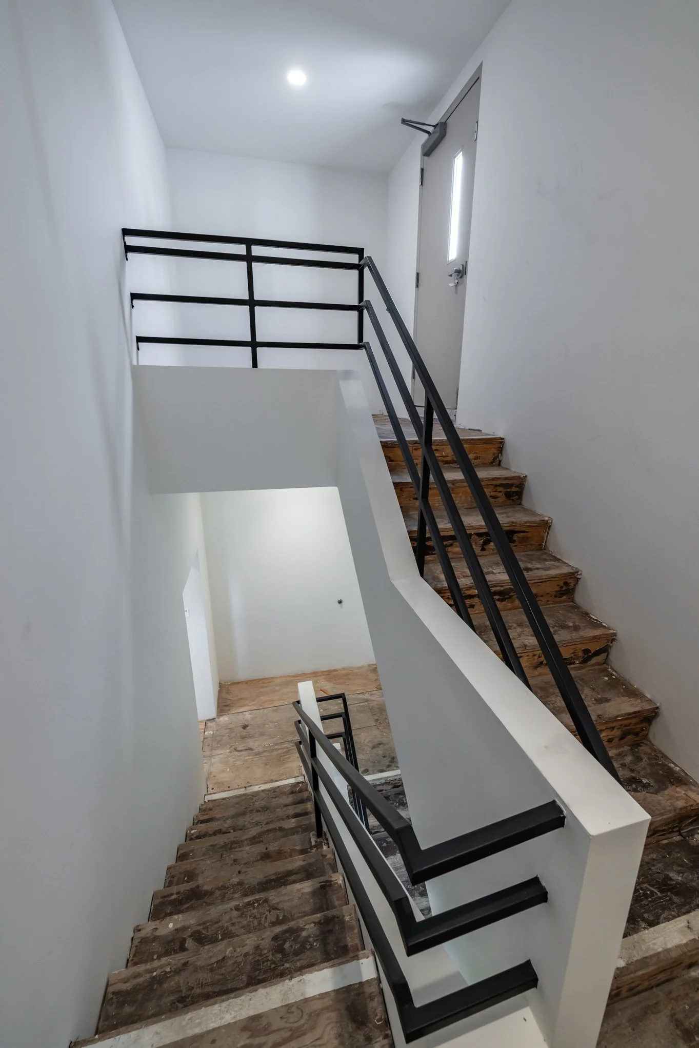 Interior view of a stairwell with unfinished wooden steps, white walls, and black metal railings leading up and down to different floors.