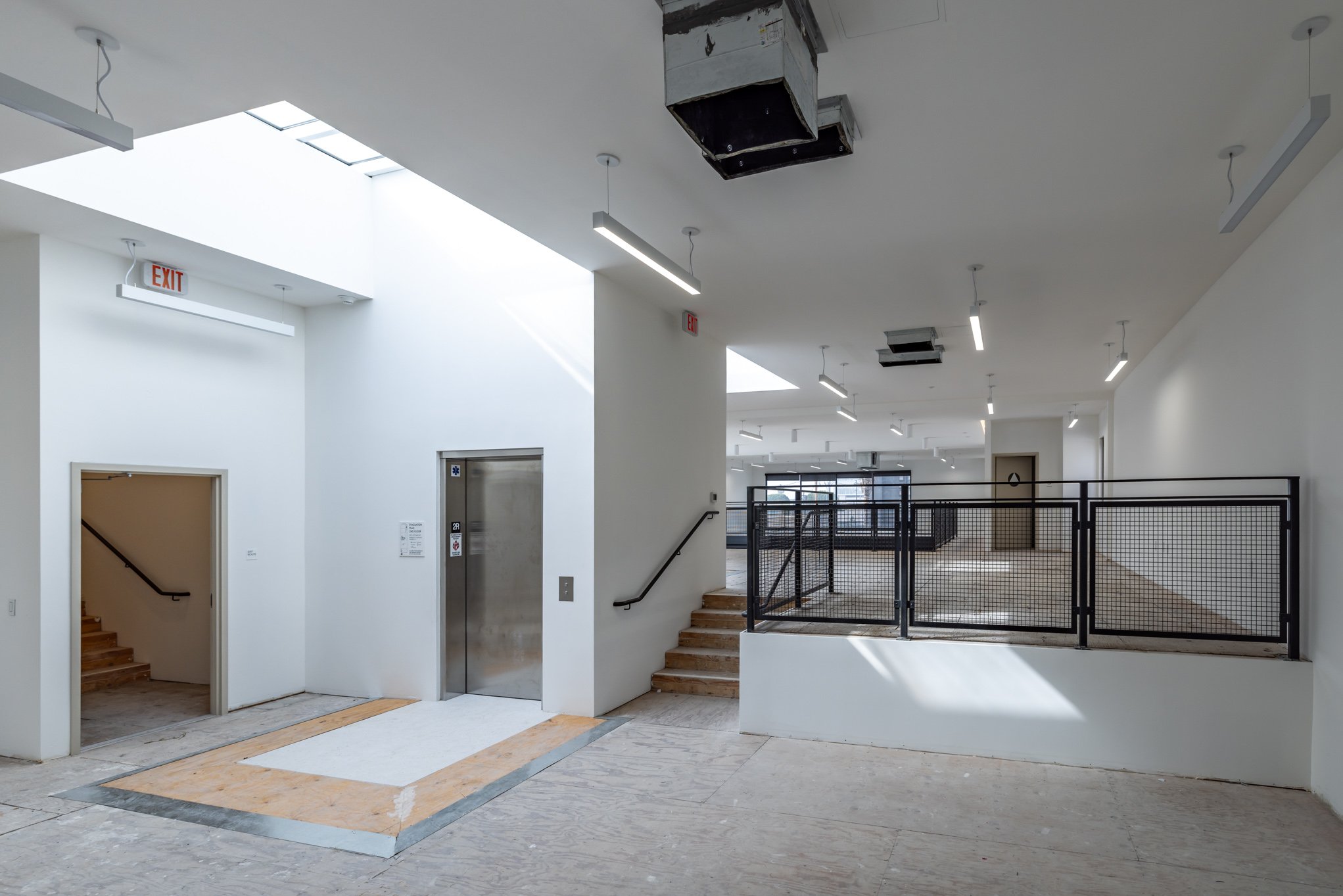 Empty indoor space with white walls, ceiling-mounted lights, an elevator, a staircase, and unfinished flooring in a building under construction or renovation.