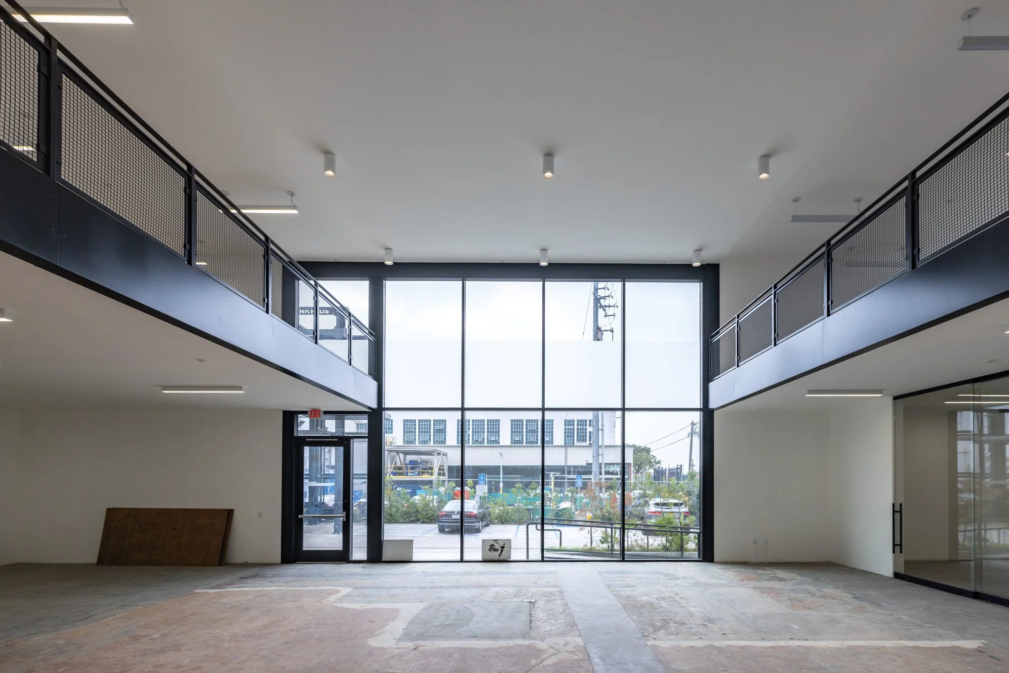 Empty interior space with large glass windows and door, second-floor balcony with black metal railing, construction materials on the floor, and cars outside.