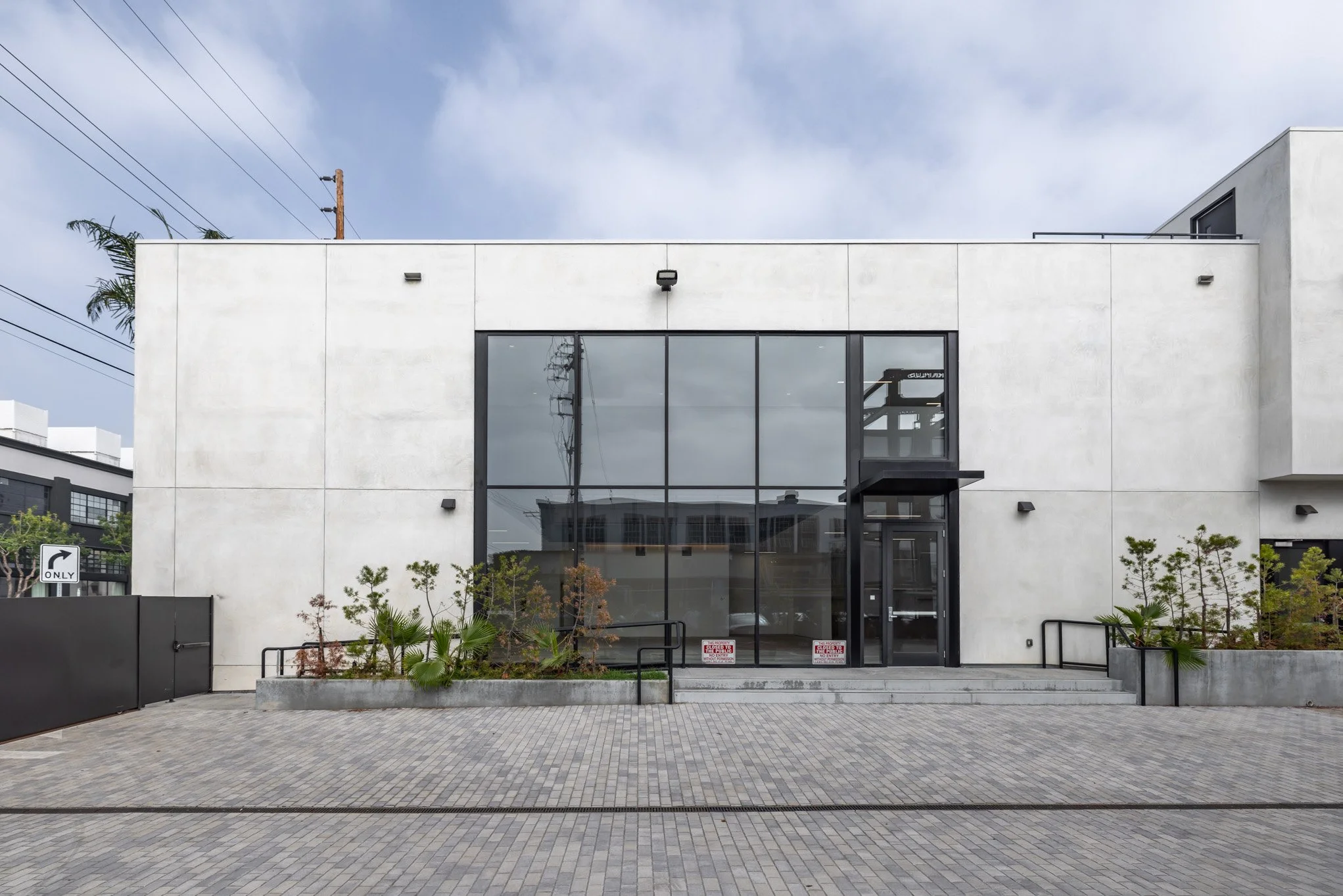 Front view of a modern, minimalist building with large glass windows, concrete exterior, small landscaped plants, and a paved entrance.