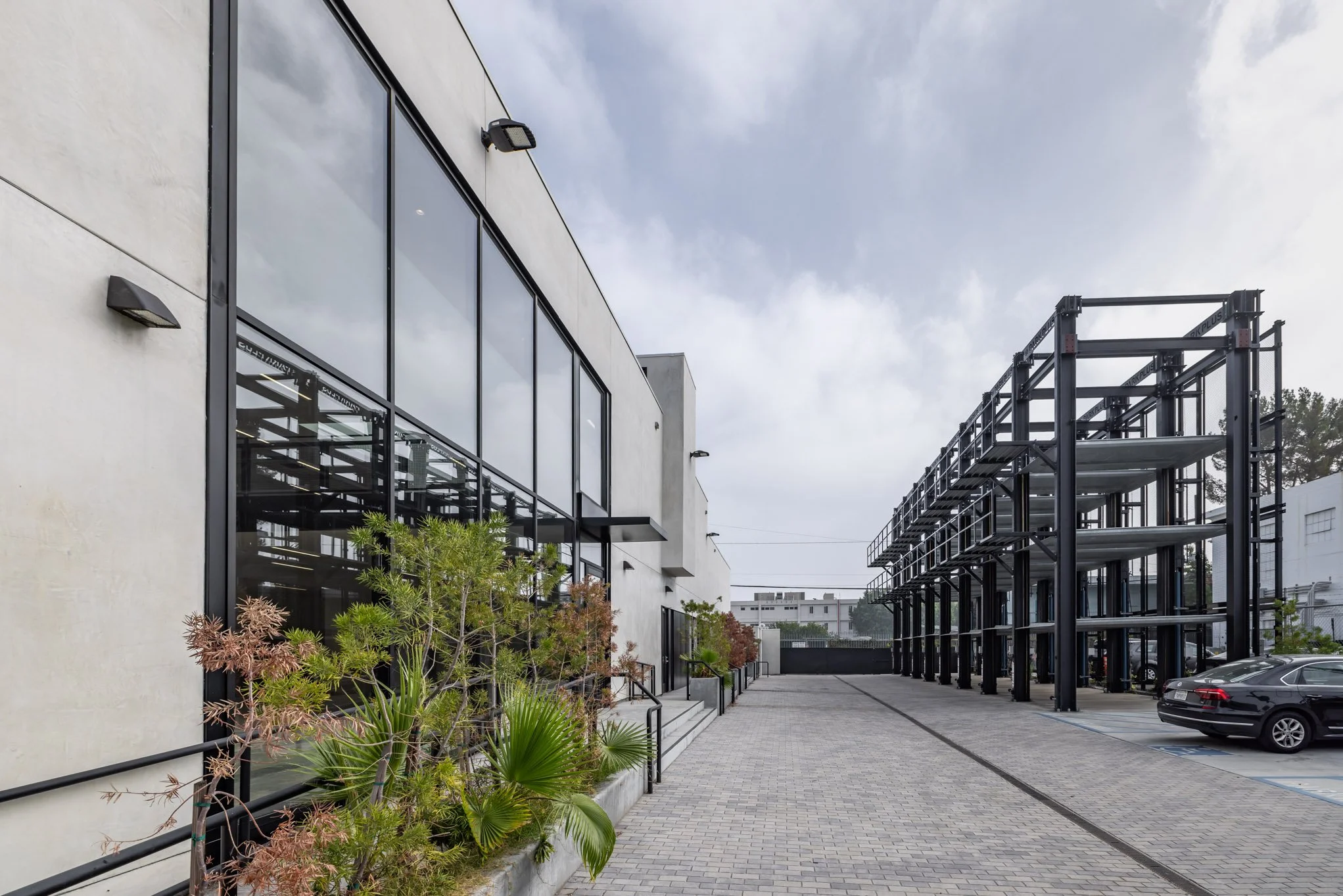A modern building with large glass windows, a landscaped walkway with plants, and an adjacent black steel parking structure with cars parked underneath.