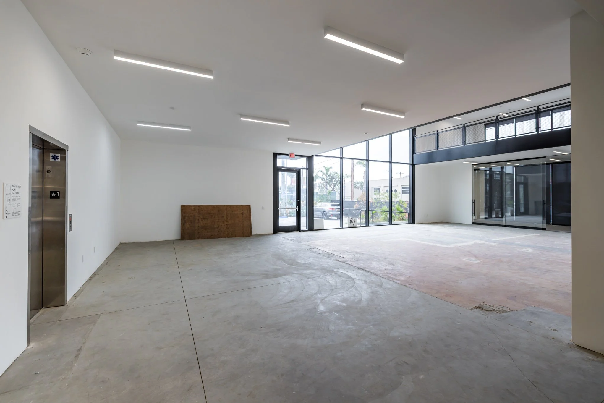 Empty commercial or retail space with large glass windows and front entrance, white walls, concrete floor, and ceiling lights.