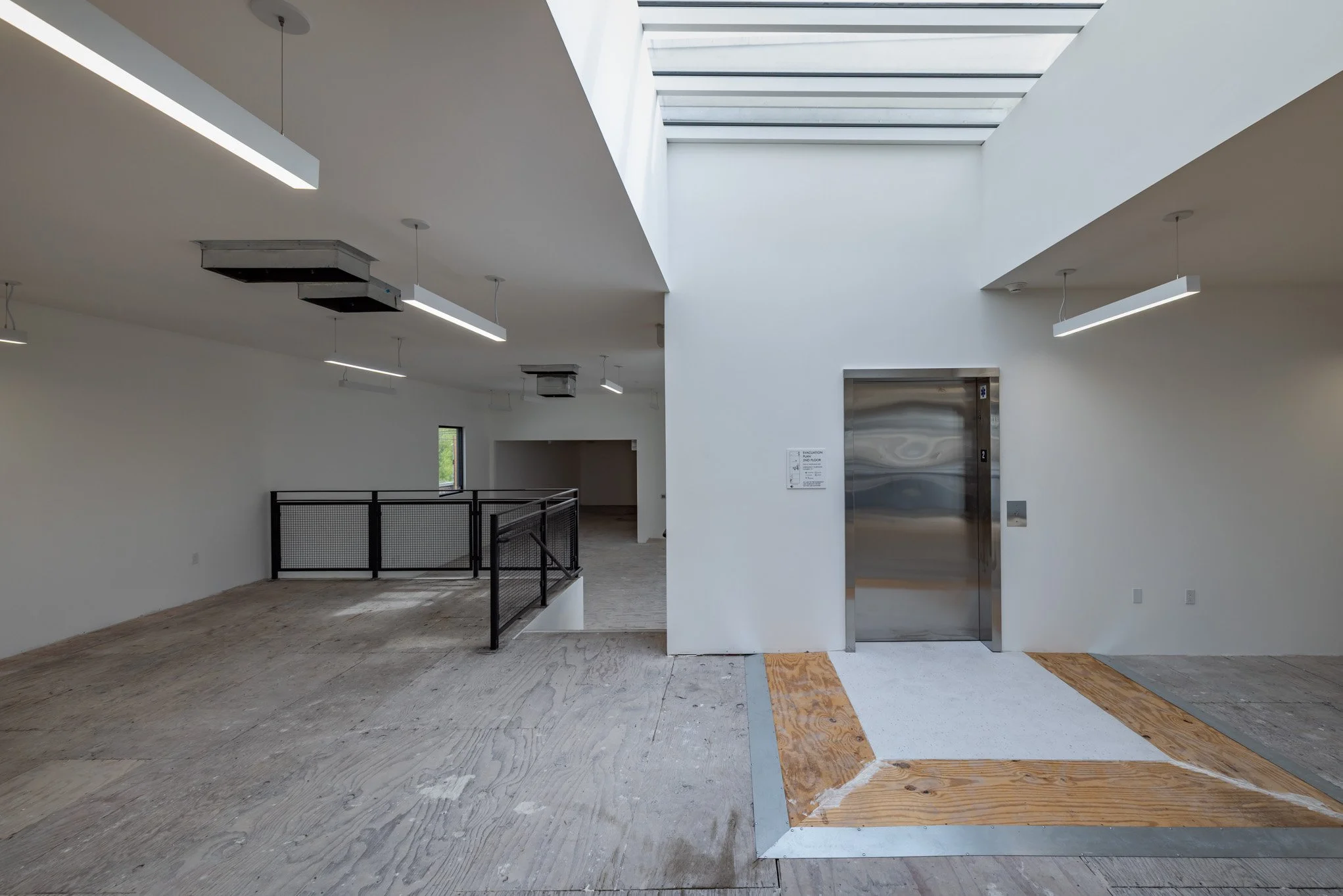 Interior space with white walls, a skylight ceiling, and an elevator. The space is under construction or renovation, with unfinished flooring and some panels missing. There are black railings and suspended lighting fixtures.