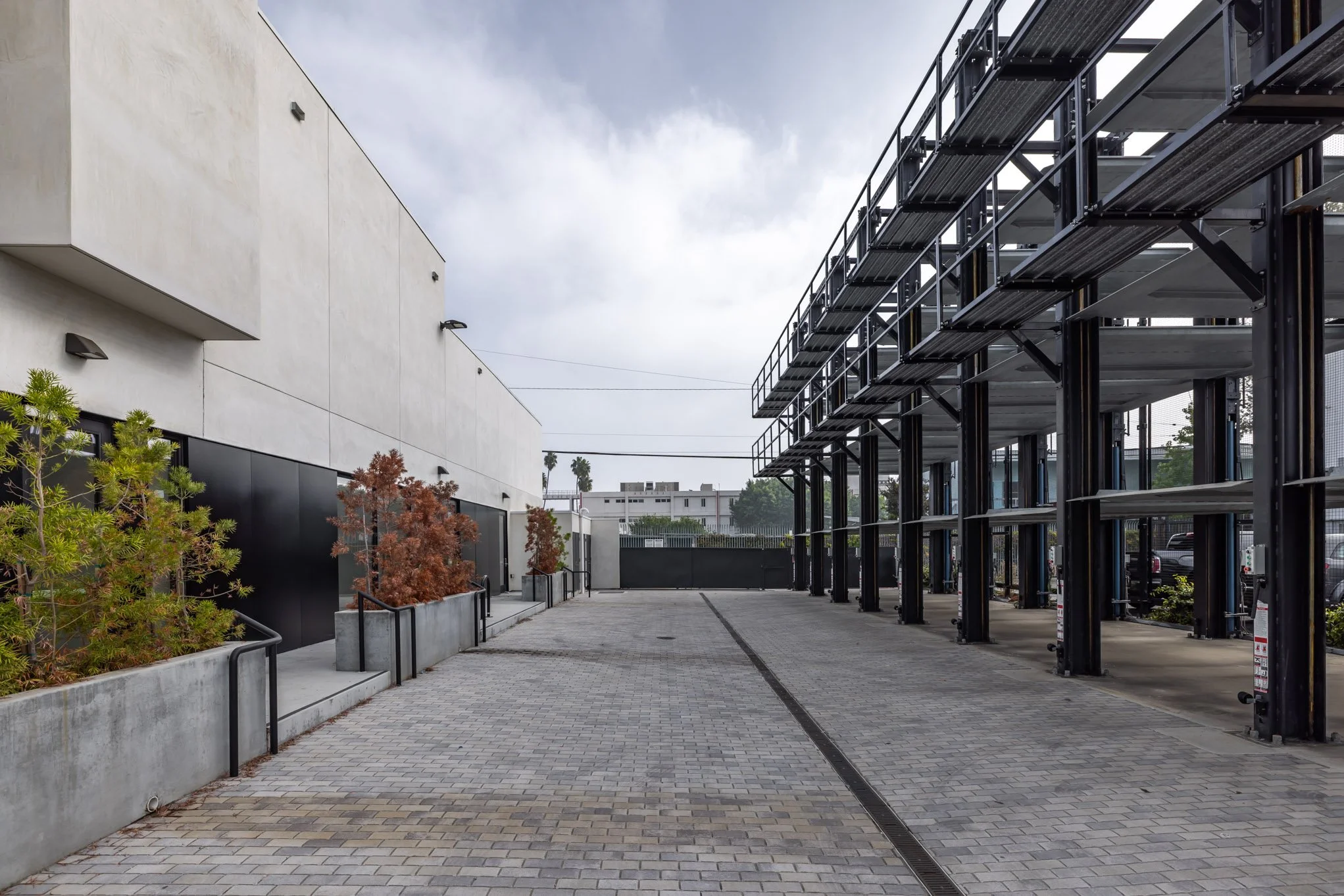 Empty outdoor space with a modern building on the left and a parking structure on the right, paved walkway, some young trees in planters, and cloudy sky.