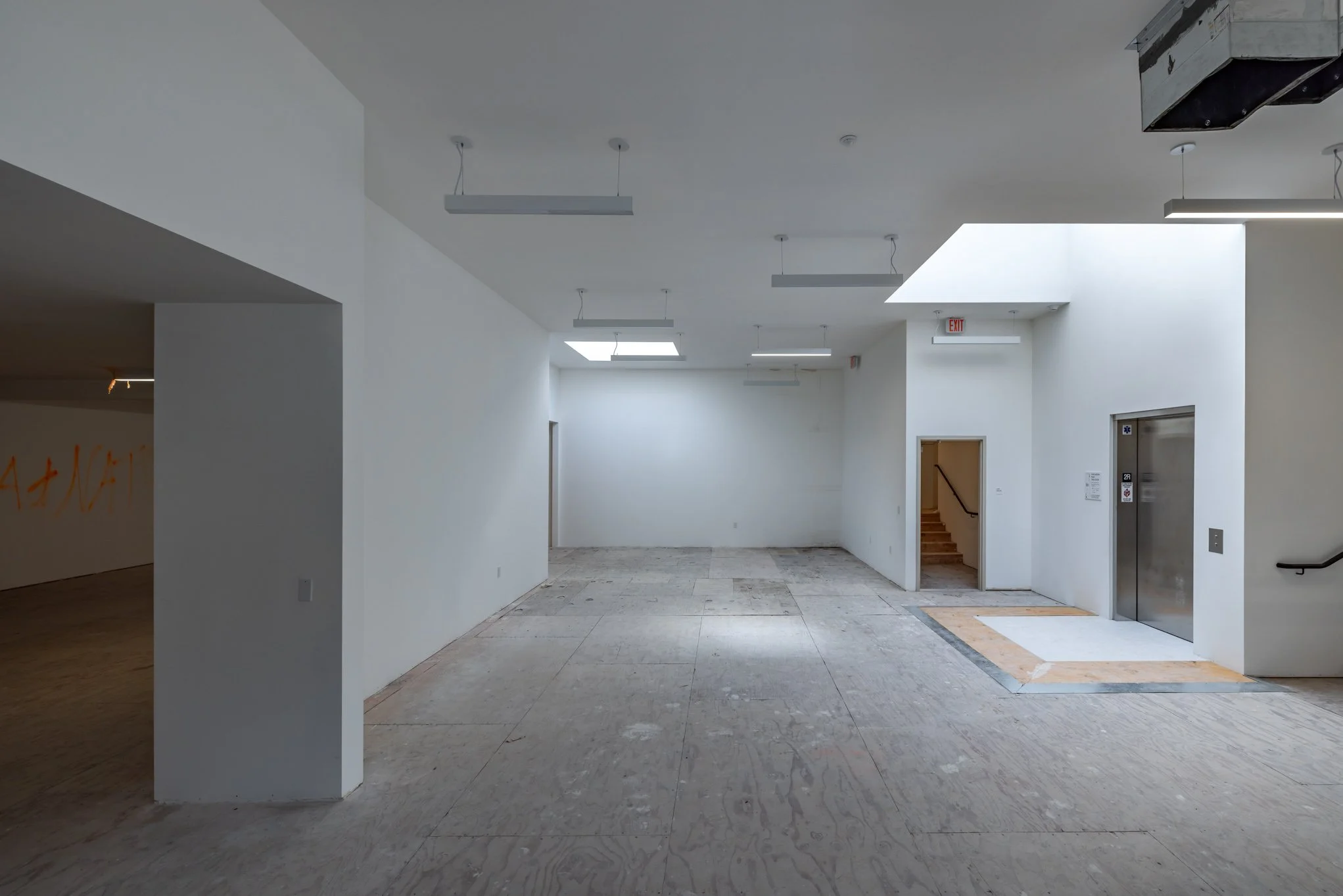 Interior of a building under construction with white walls, unfinished floors, and an elevator. An open doorway leads to stairs, and there are lighting fixtures hanging from the ceiling.