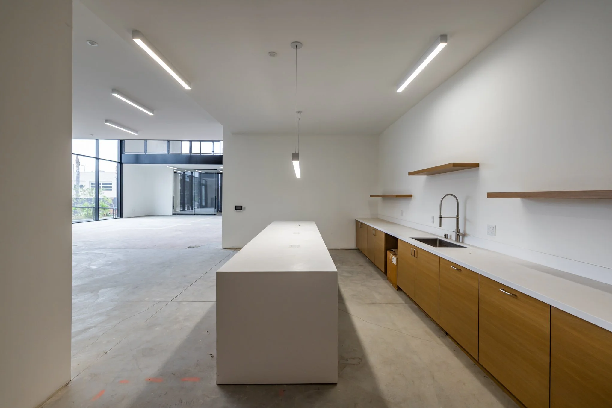 Empty modern kitchen area with wooden cabinets, white countertops, a sink, open shelves, and a white island in a spacious room with large windows and glass doors in the background.
