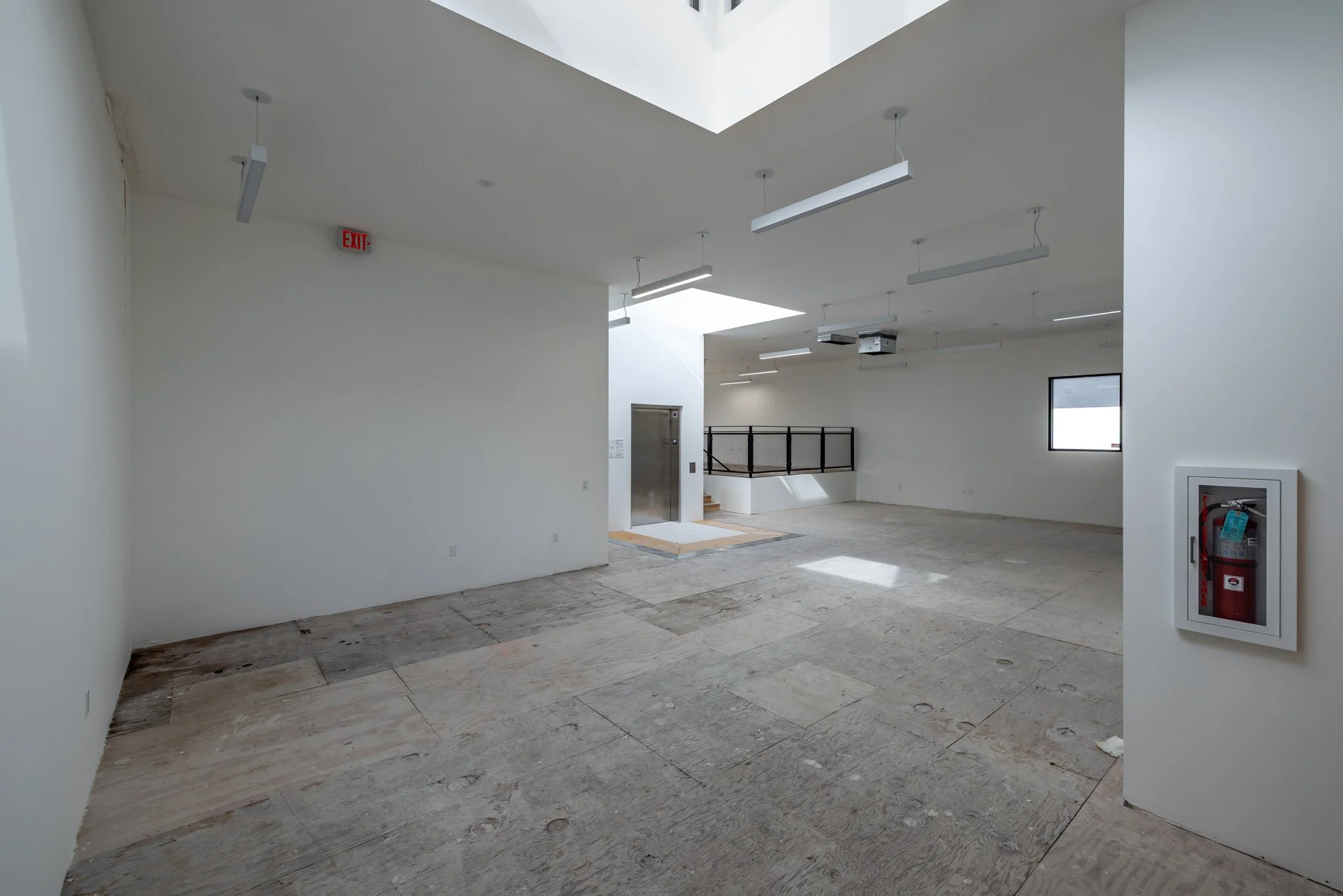 Empty interior room with white walls, unfinished wooden floor, ceiling lights, small window, and a fire extinguisher in a wall-mounted cabinet.