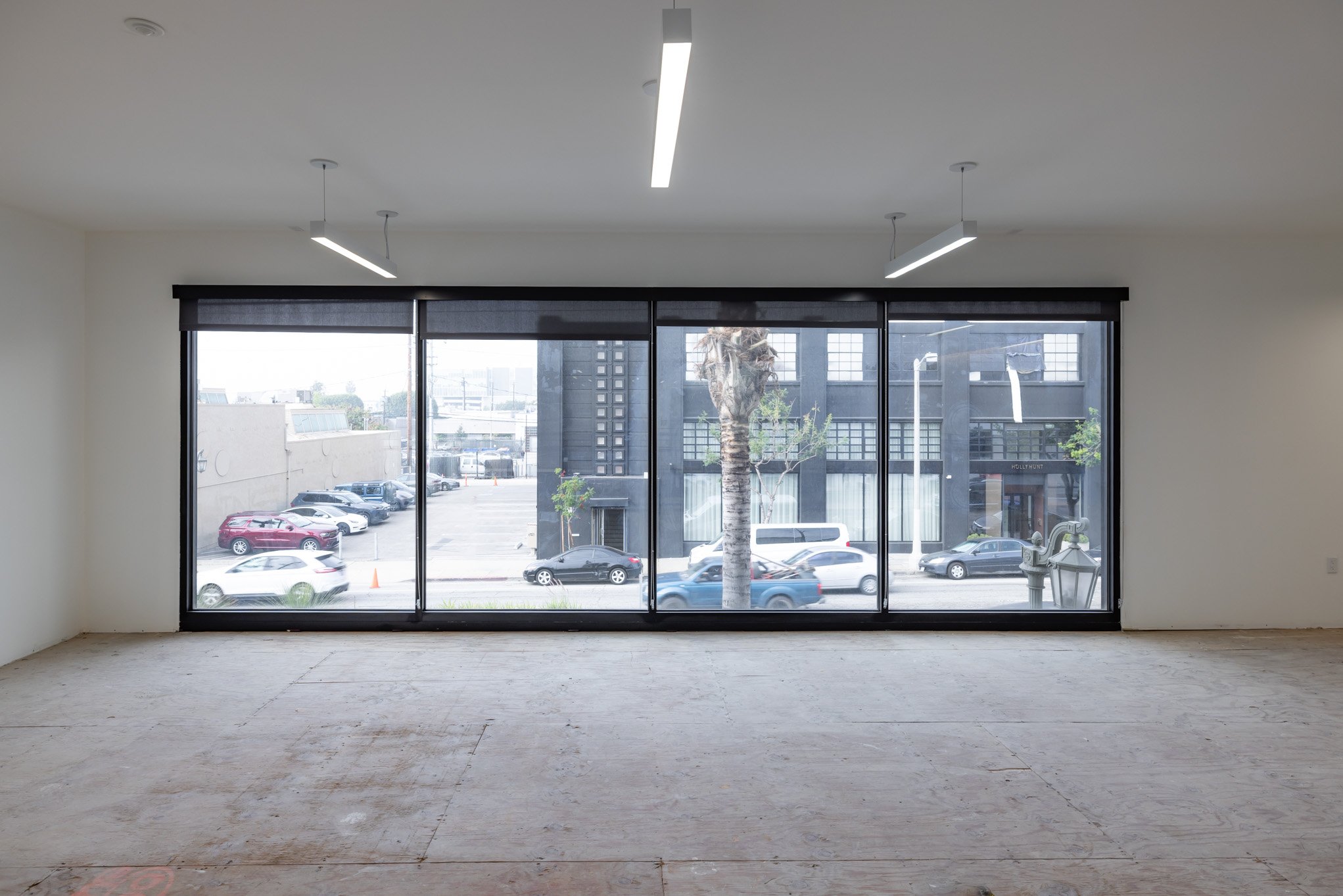 Empty room with large front window showing a parking lot and city street outside, with parked cars and a tree visible.