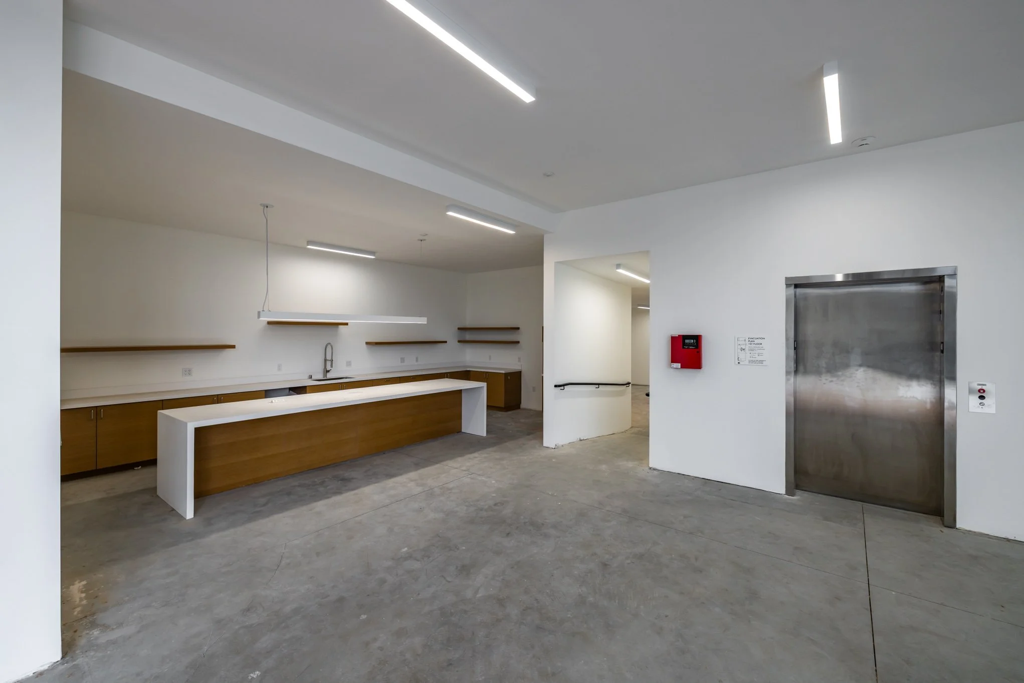 Empty modern room with white walls, concrete floor, kitchen area with wooden cabinets, white countertop, and open shelves, and an elevator door.