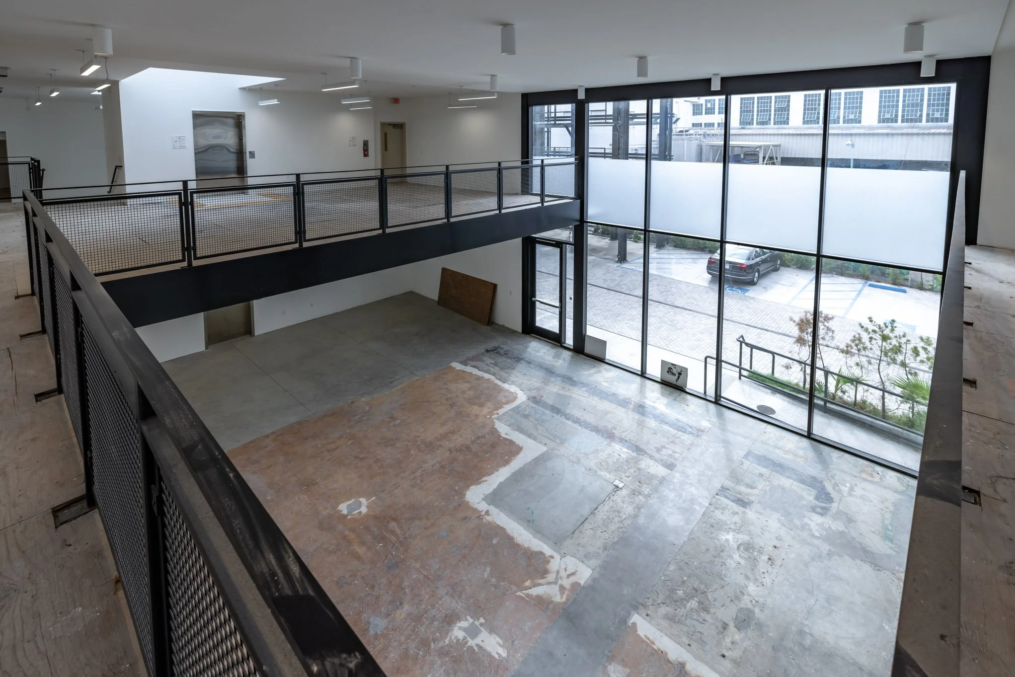 Interior of an unfinished commercial building with large glass windows, a two-level layout with metal railings, and an empty space on the ground floor with visible patches of concrete and flooring material.