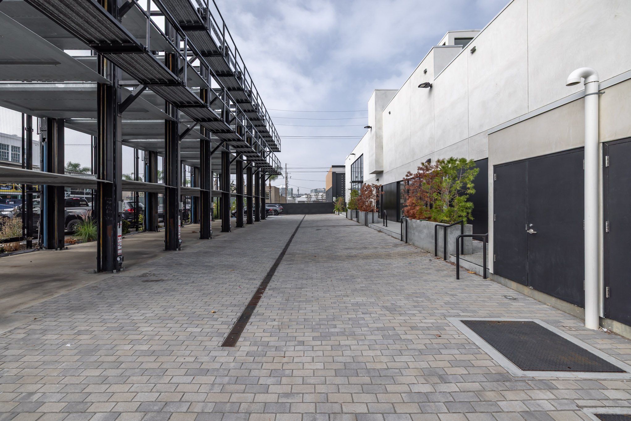 Empty urban sidewalk bordered by a modern building on the right with plants, and a parking area on the left with metal parking structures.