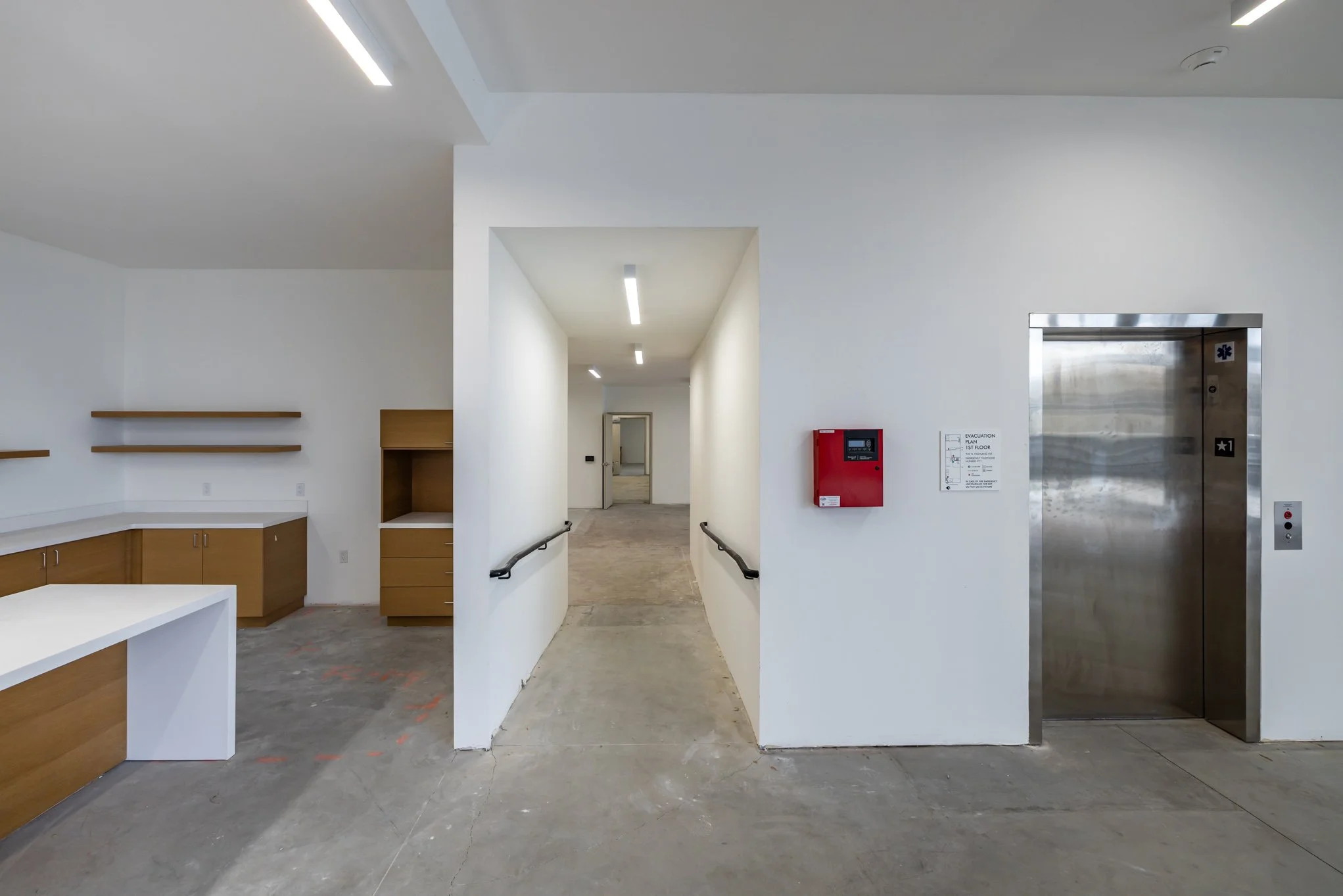 Empty hospital corridor with white walls, wooden cabinets, and an elevator on the right. Hallway has handrails, a red fire alarm box, and an evacuation plan on the wall.