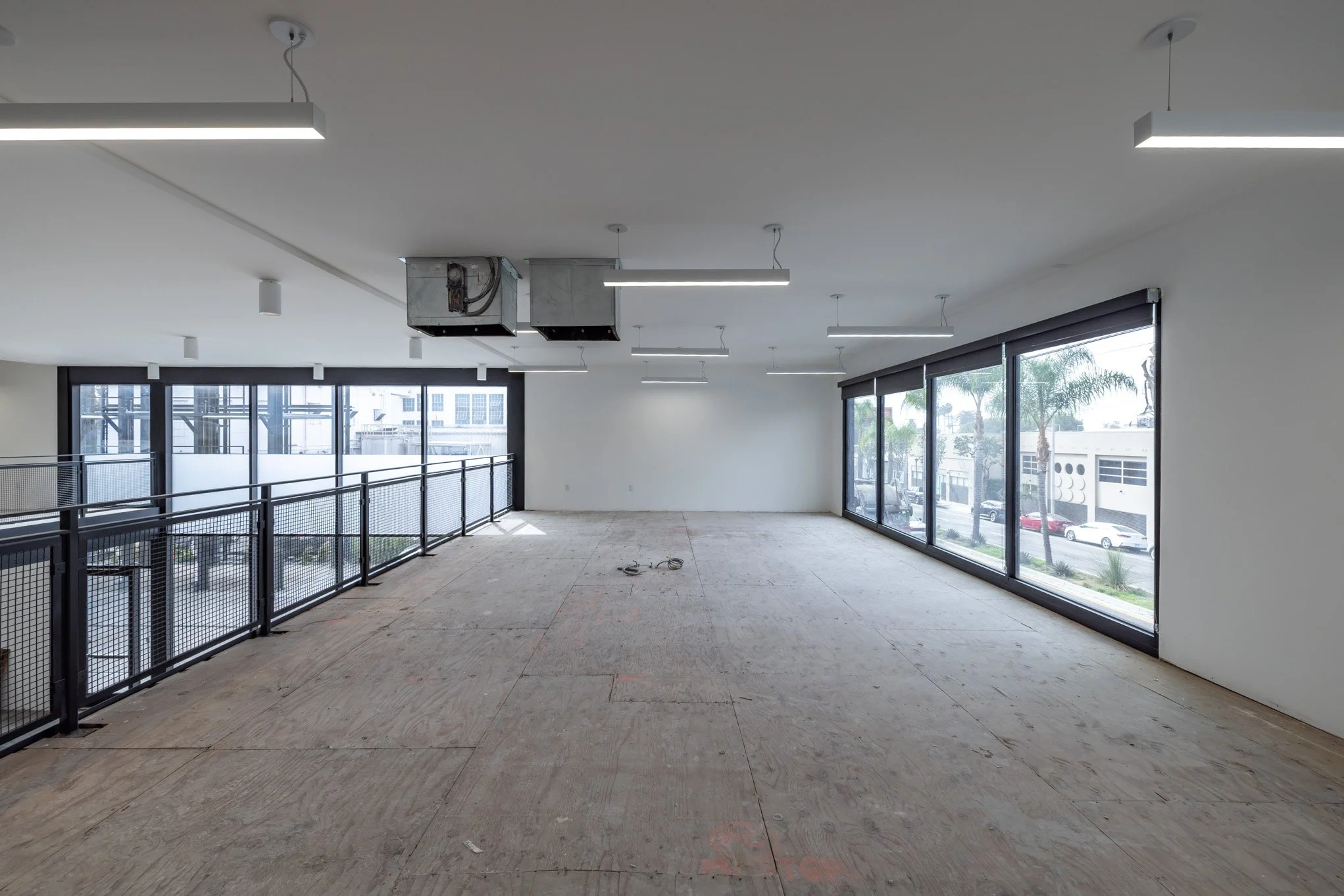Empty commercial space with large windows, unfinished flooring, and ceiling lights.