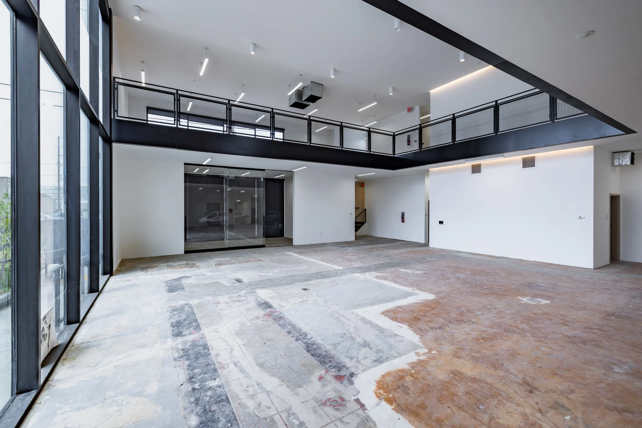 Empty interior space with large glass windows on the left, white walls, a second-floor black railing on the upper level, exposed ductwork, and a partially unfinished floor.