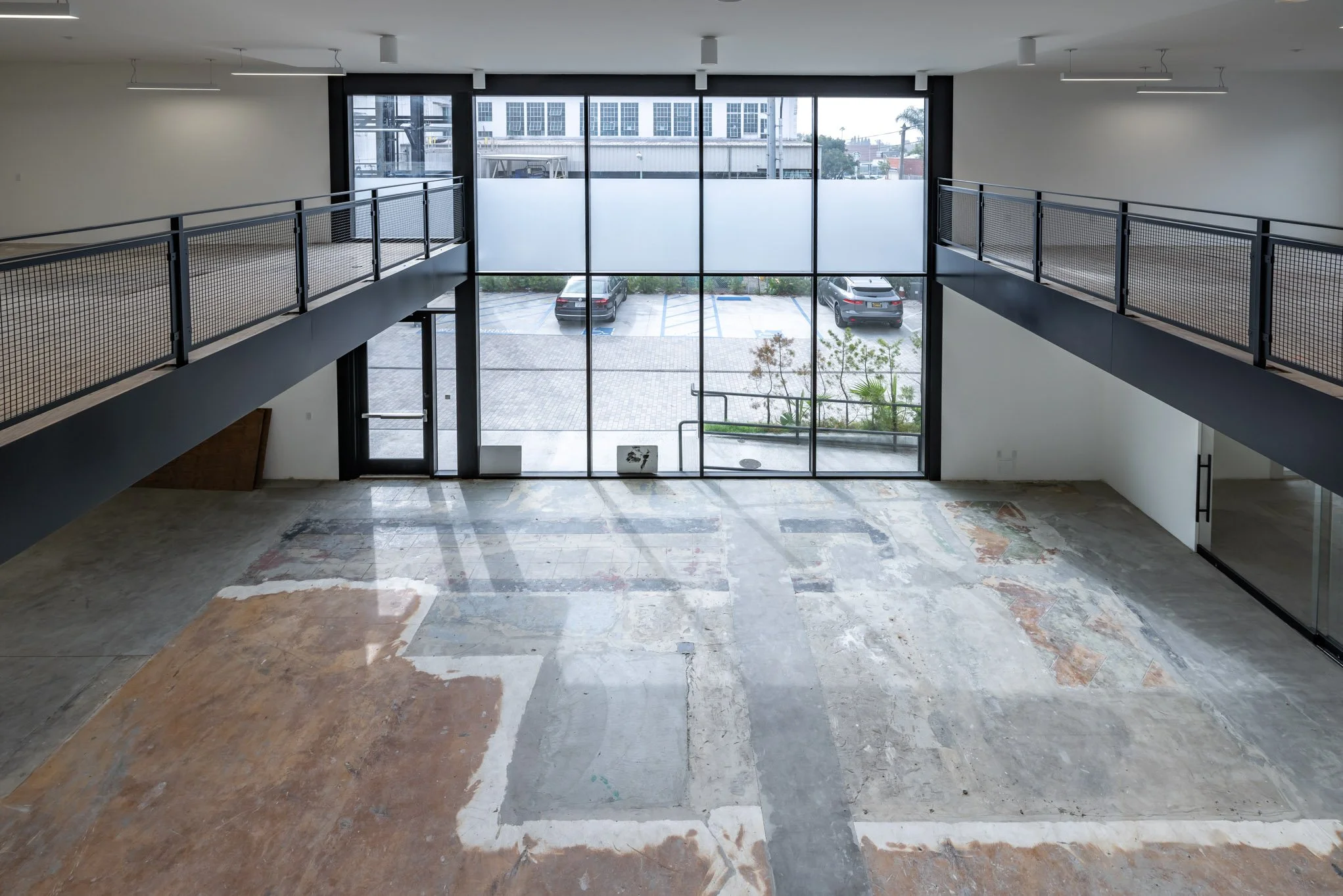 Empty commercial space with unfinished flooring, large glass windows, and a balcony facing parking lot.